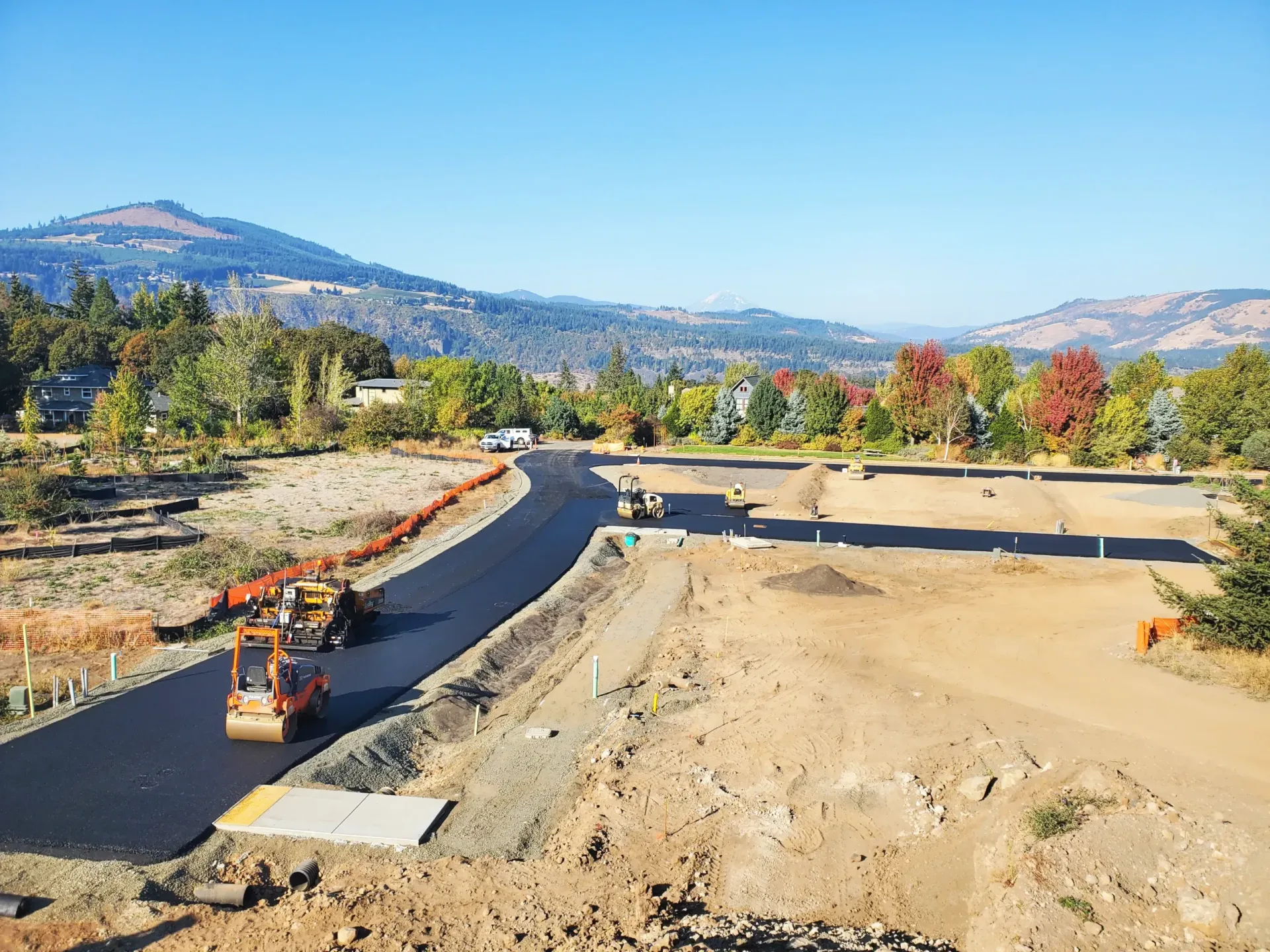 A construction site with a road being paved by rollers, set against a background of trees and mountains under a blue sky.