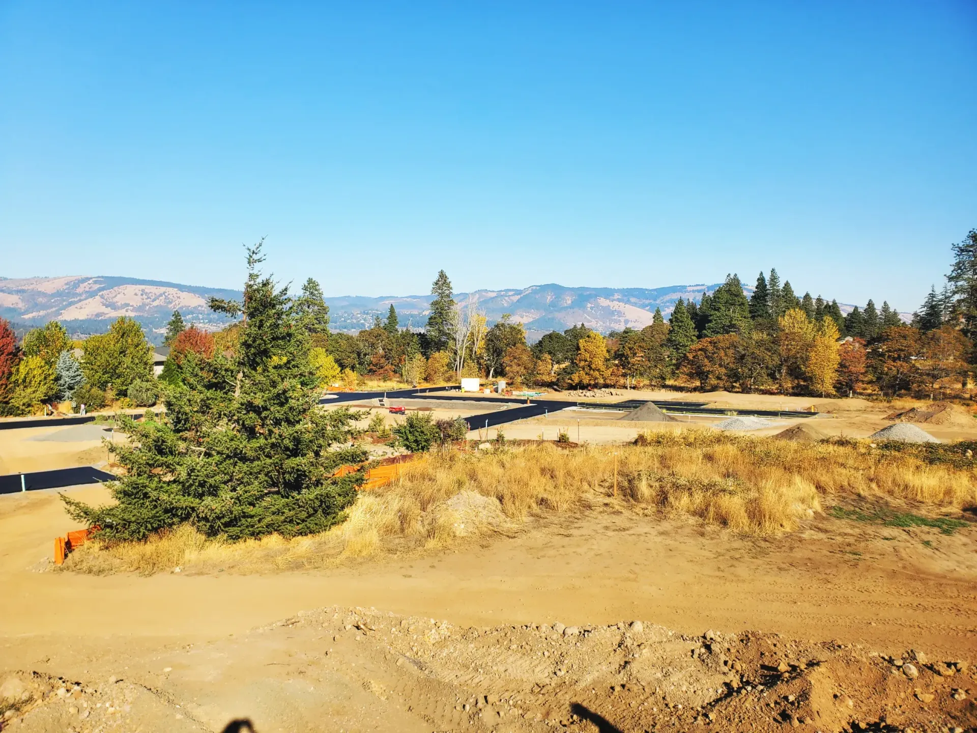 An expansive construction site with dirt mounds, a gravel road, and surrounding autumn-colored trees under a blue sky.