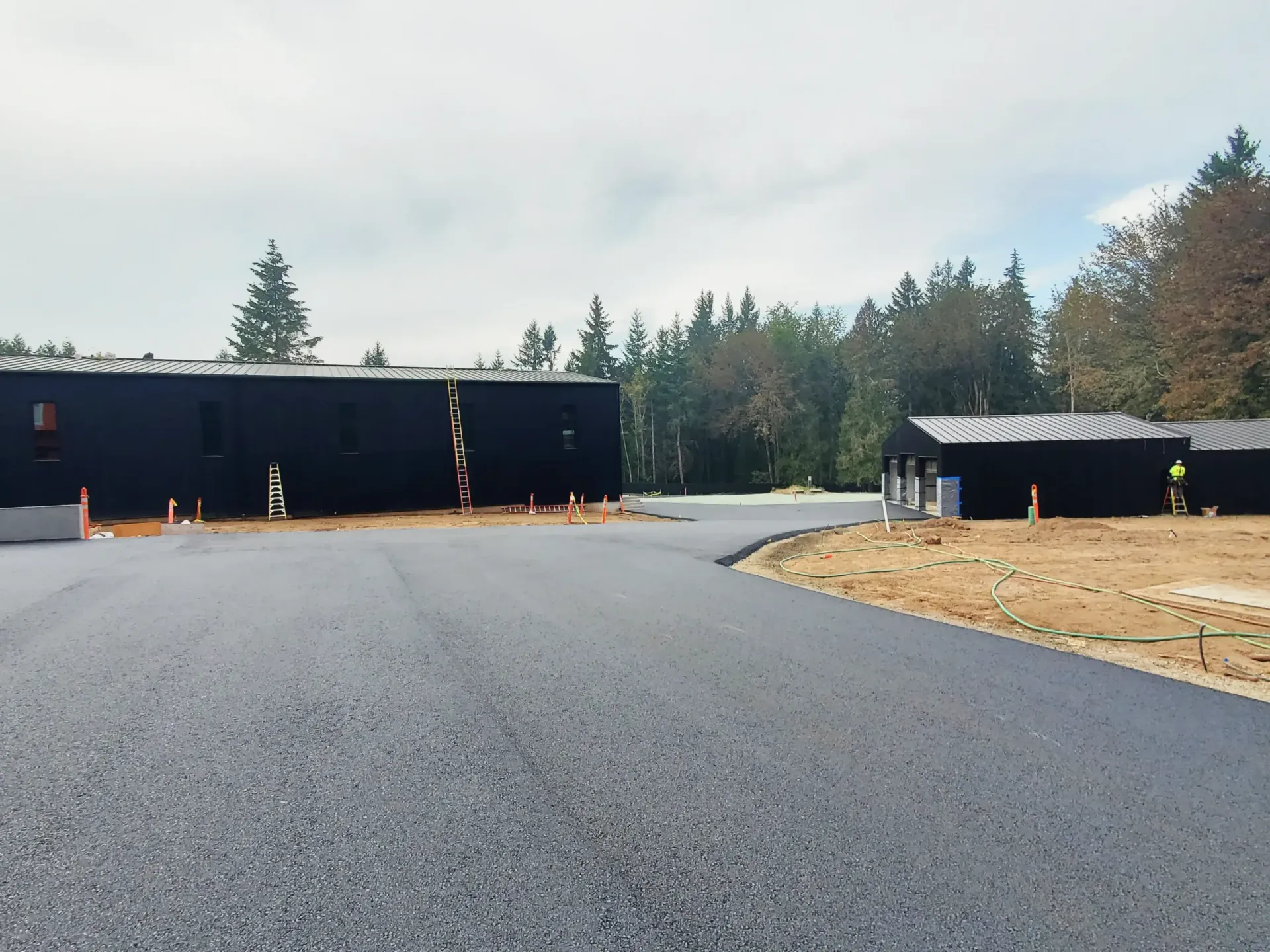 Freshly paved asphalt driveway leading between two dark, modern buildings surrounded by trees under a cloudy sky.