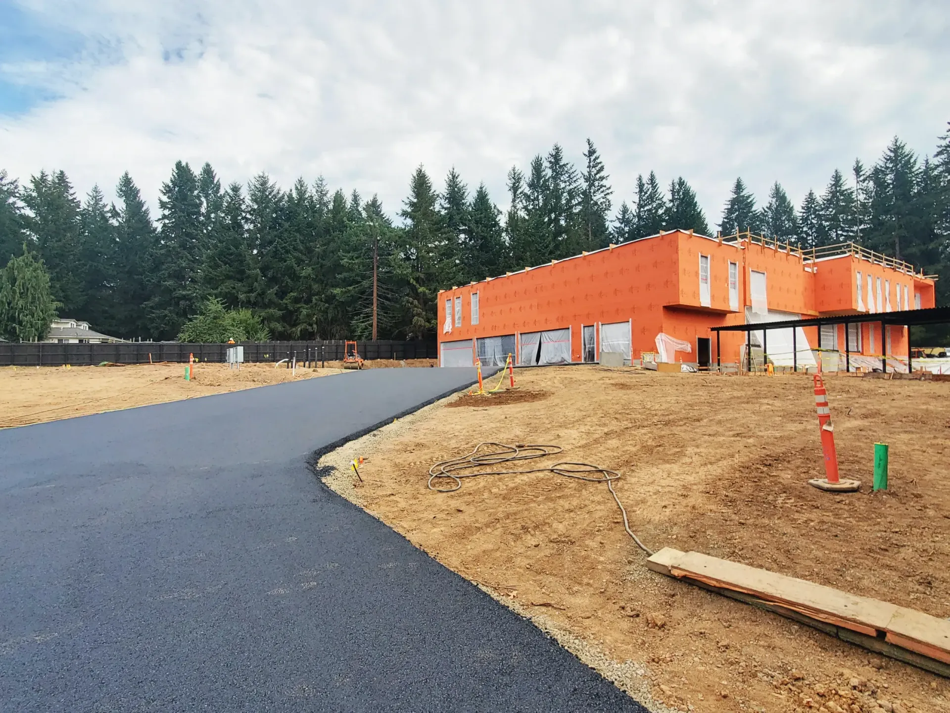 A new asphalt driveway curves toward a building under construction, featuring bright orange exterior sheathing.