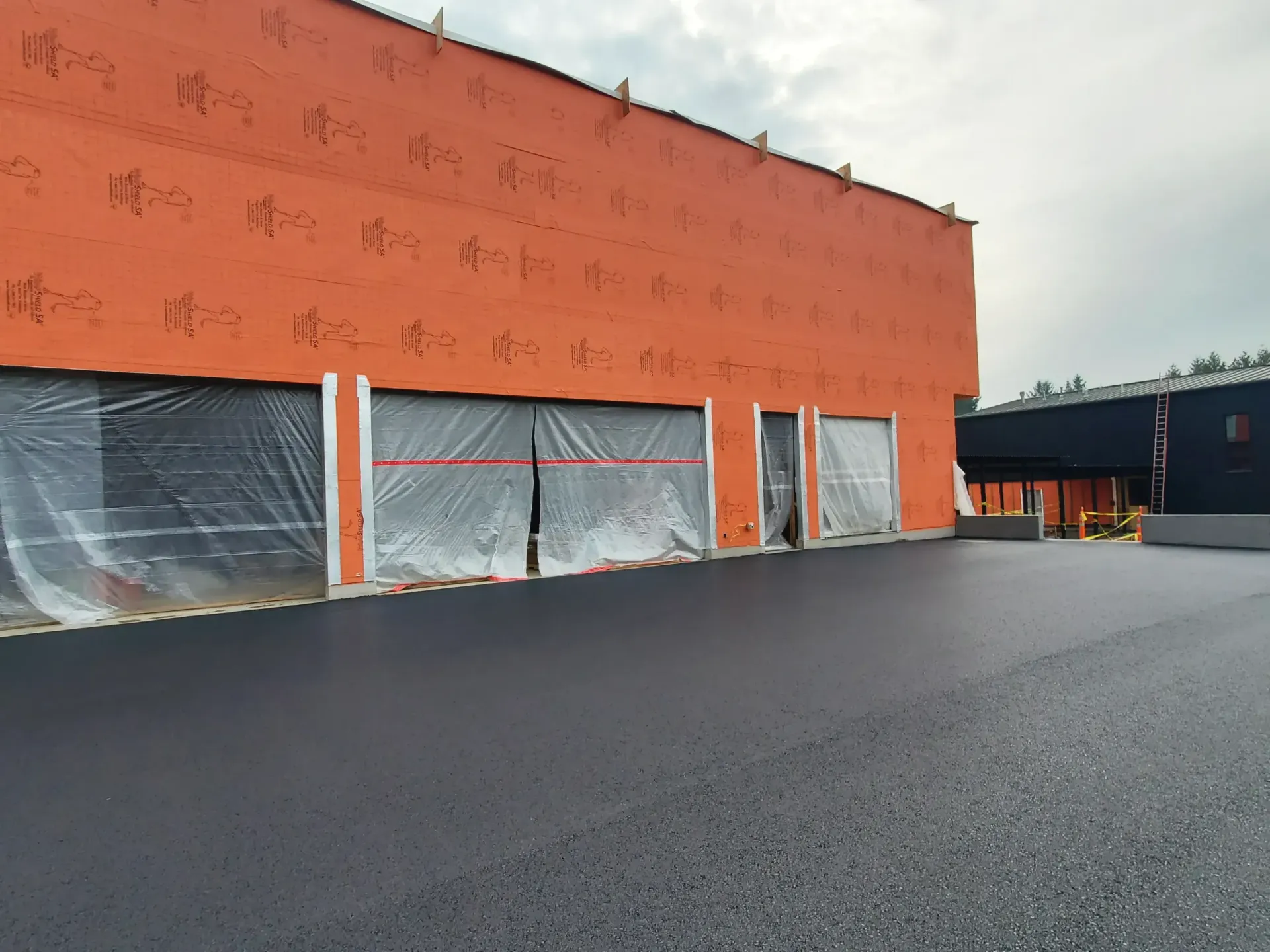 Newly paved asphalt parking lot in front of a building with orange sheathing and plastic-covered openings.