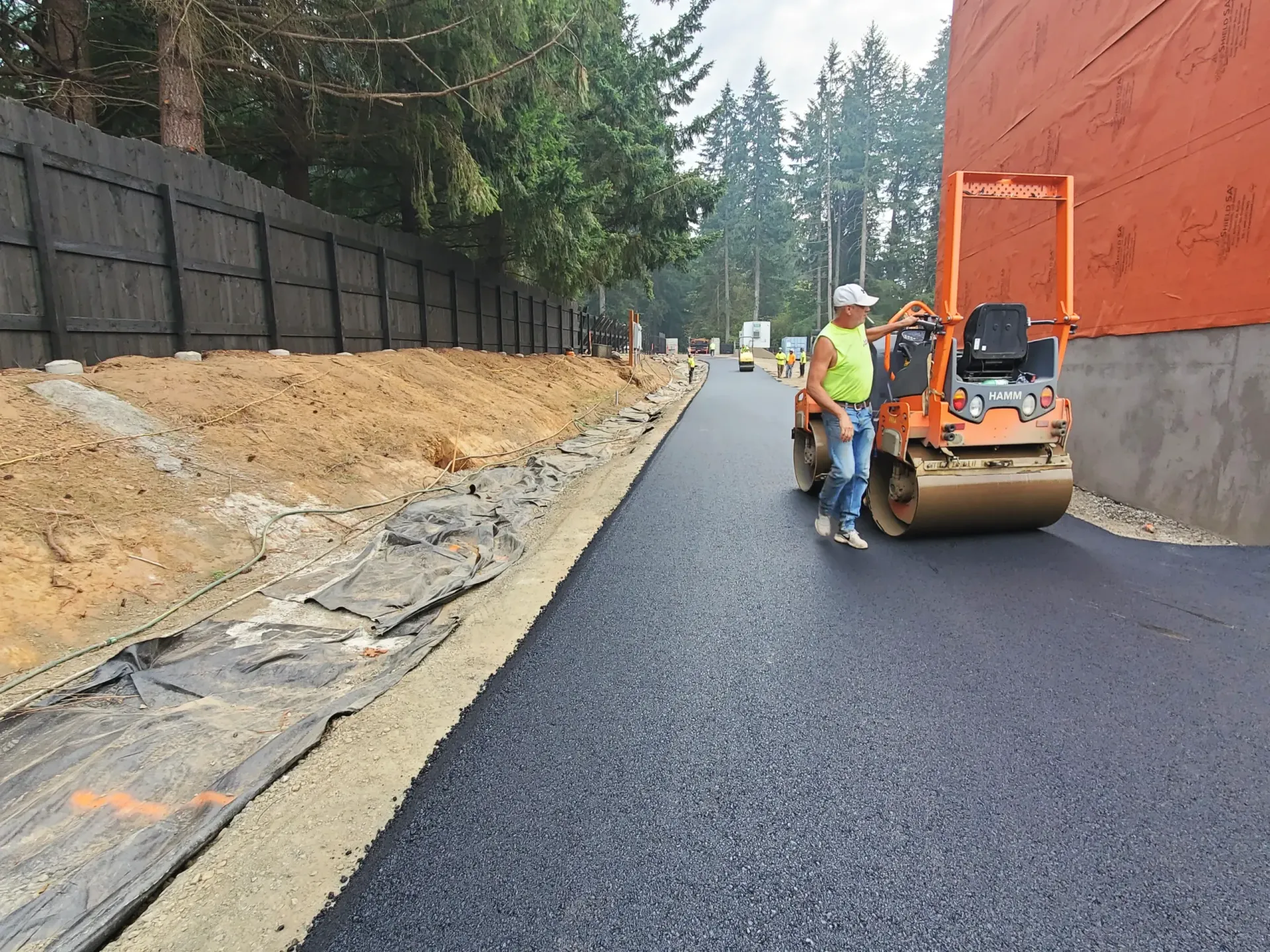 A worker stands next to an orange asphalt roller on a newly paved road bordered by a dirt embankment and a wooden fence.