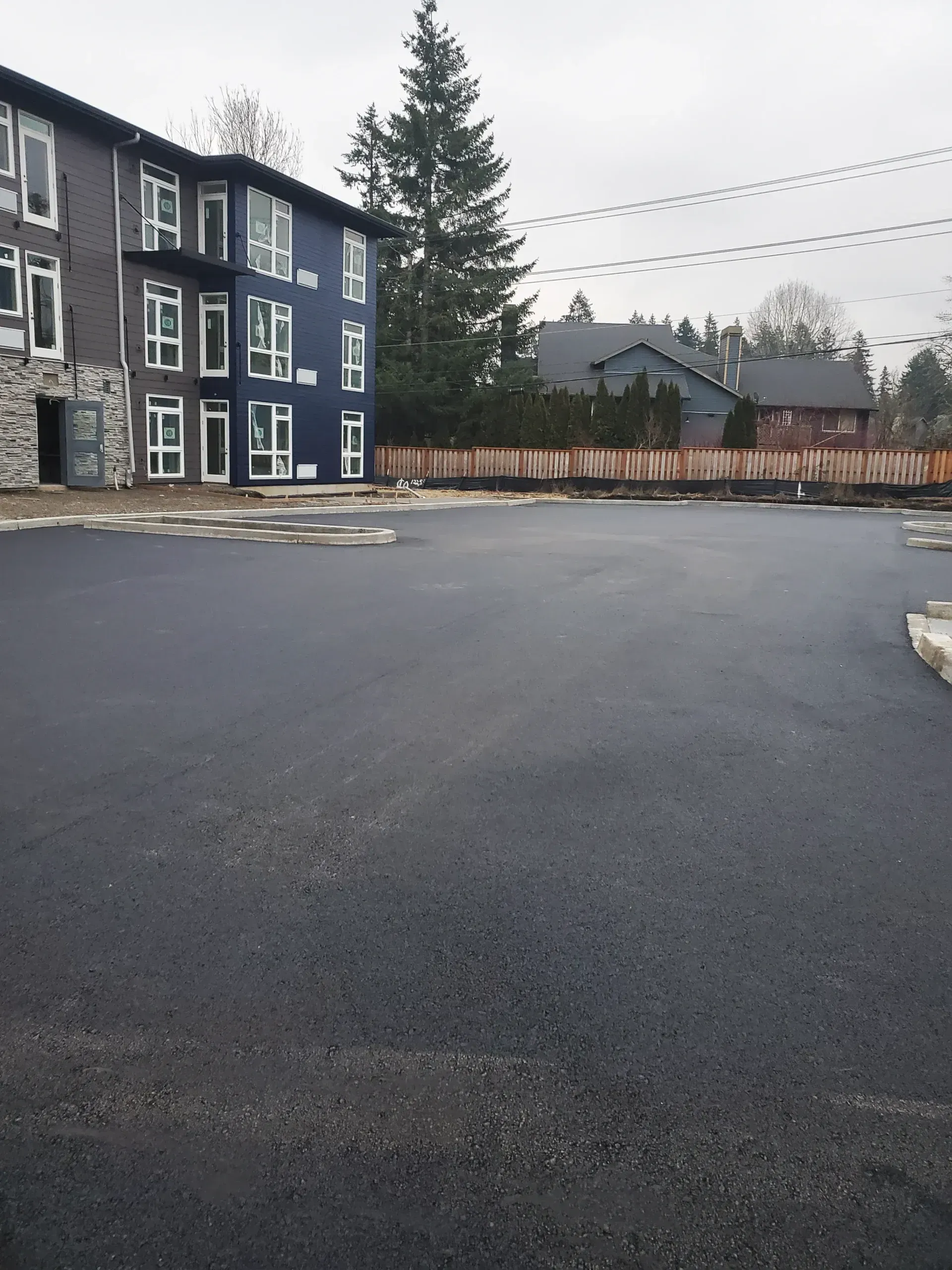 A large, newly paved dark asphalt parking lot sits in front of a blue and grey three-story apartment building.