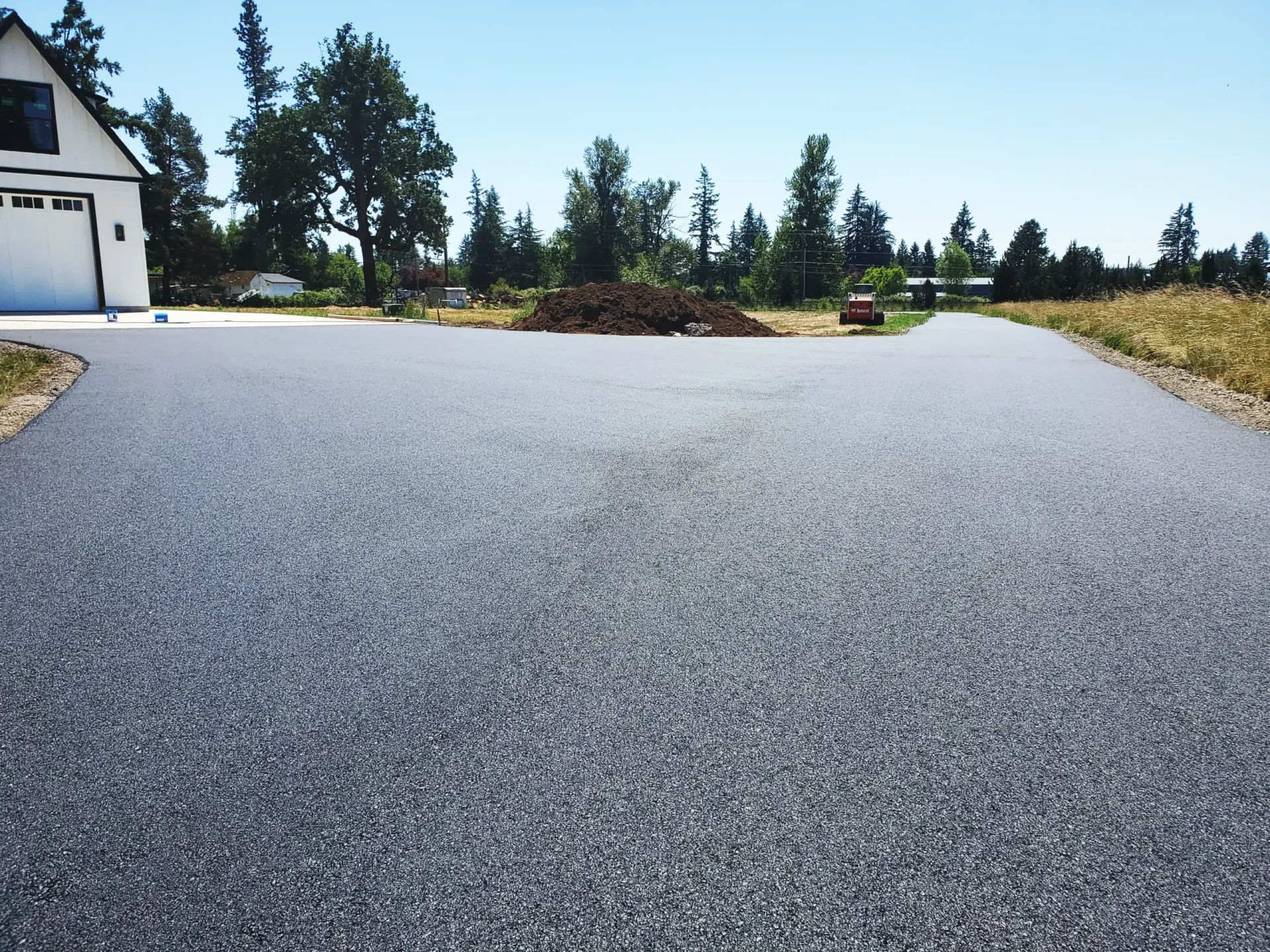 A freshly paved asphalt driveway leading toward a house and open field under a clear blue sky.