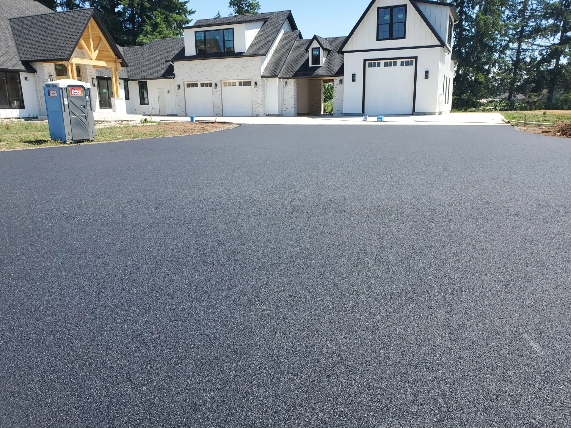 A freshly paved black asphalt driveway leads toward a multi-car garage of a modern, newly constructed suburban home.