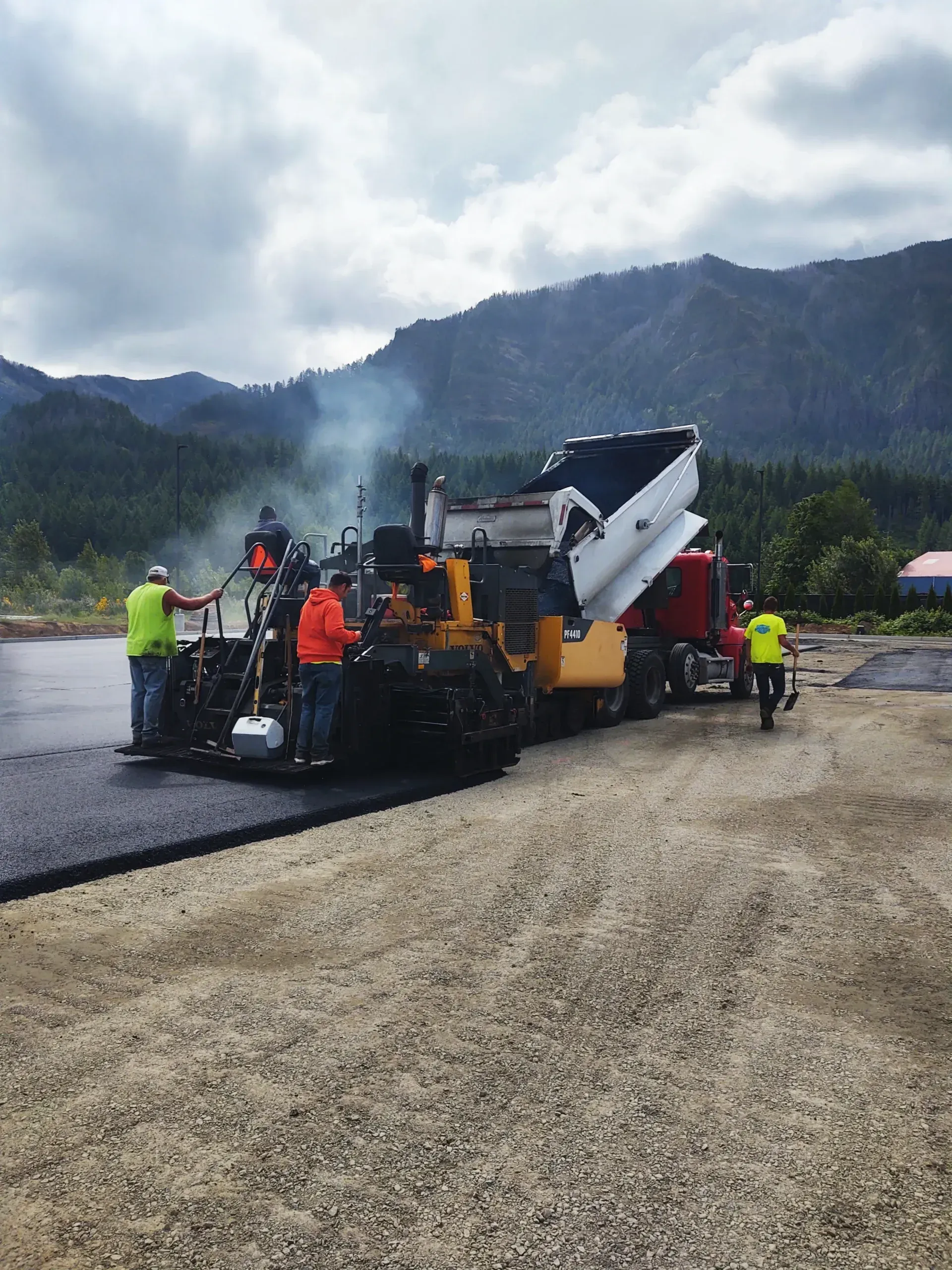A crew operates a paving machine on a gravel road, with a dump truck and mountains in the background.