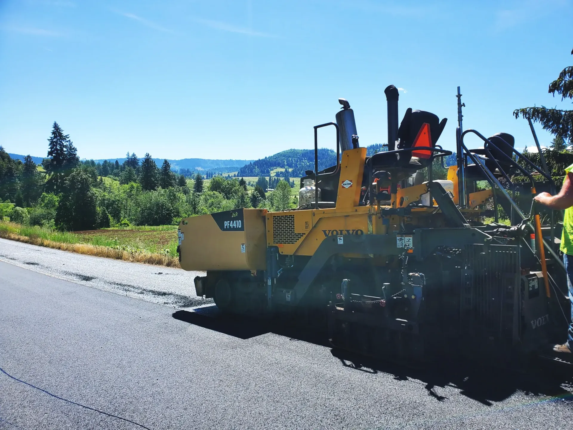 A yellow asphalt paver sits on a freshly paved road under a bright blue sky with a rural, hilly landscape in the background.