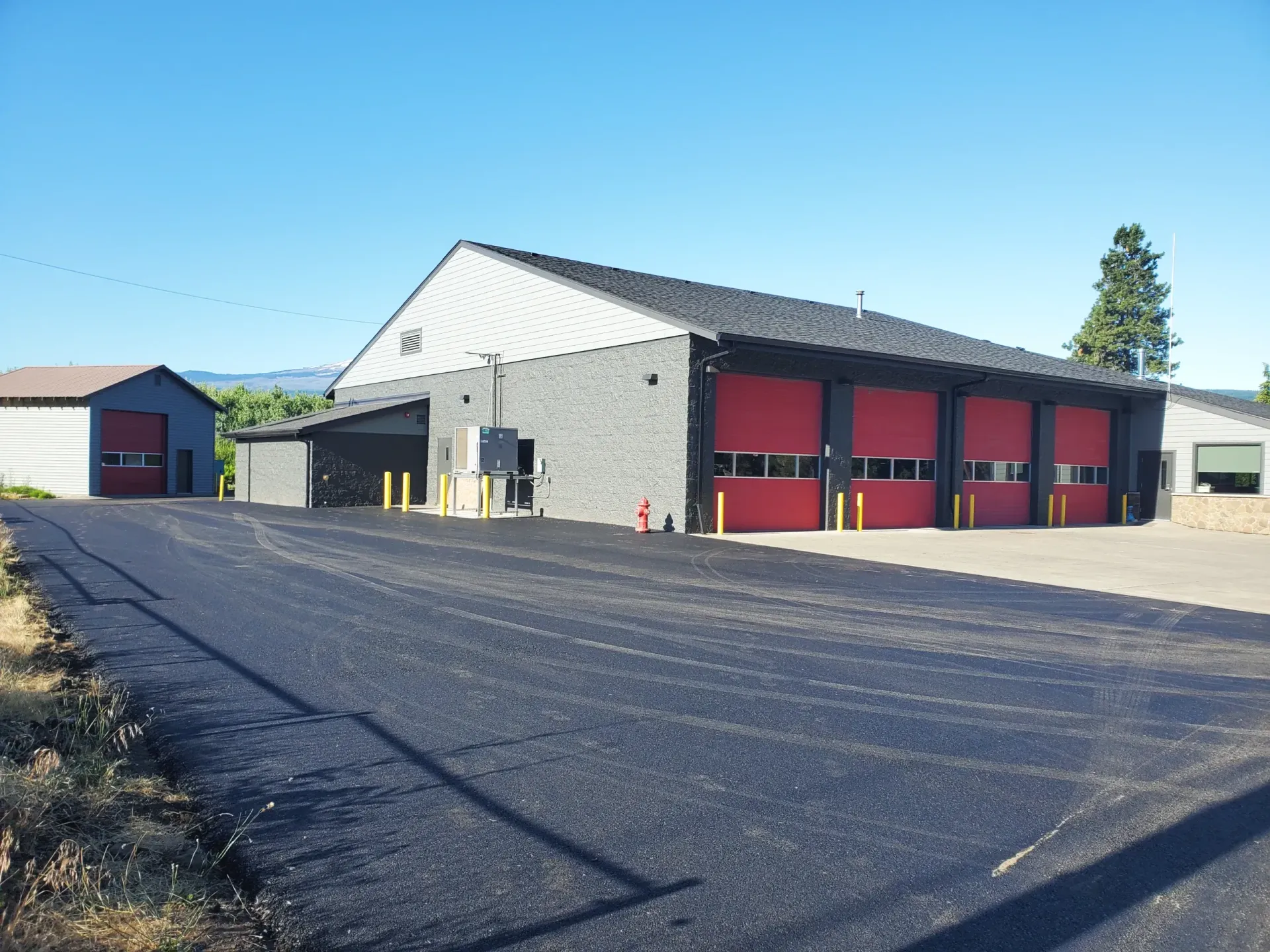 A grey building with four red bay doors and a freshly paved black asphalt driveway under a clear blue sky.