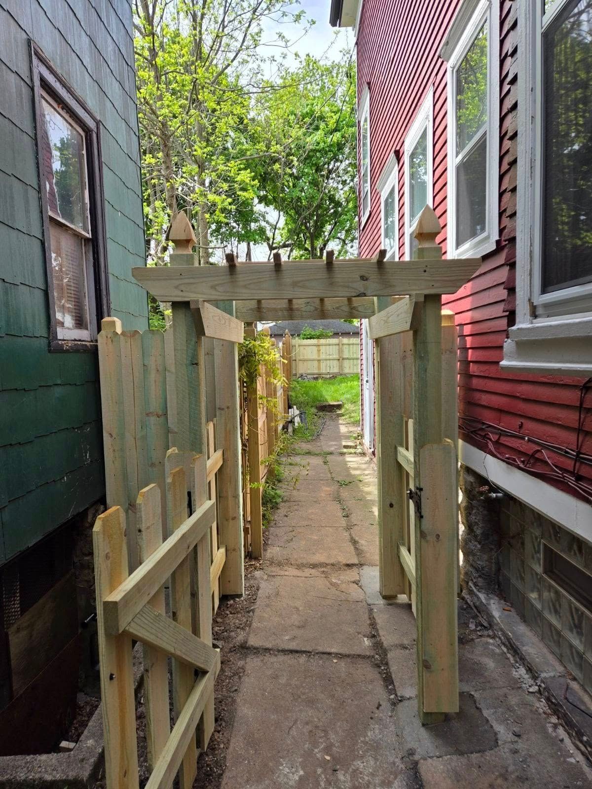 Narrow pathway between houses with a wooden gate, fence, and trellis.
