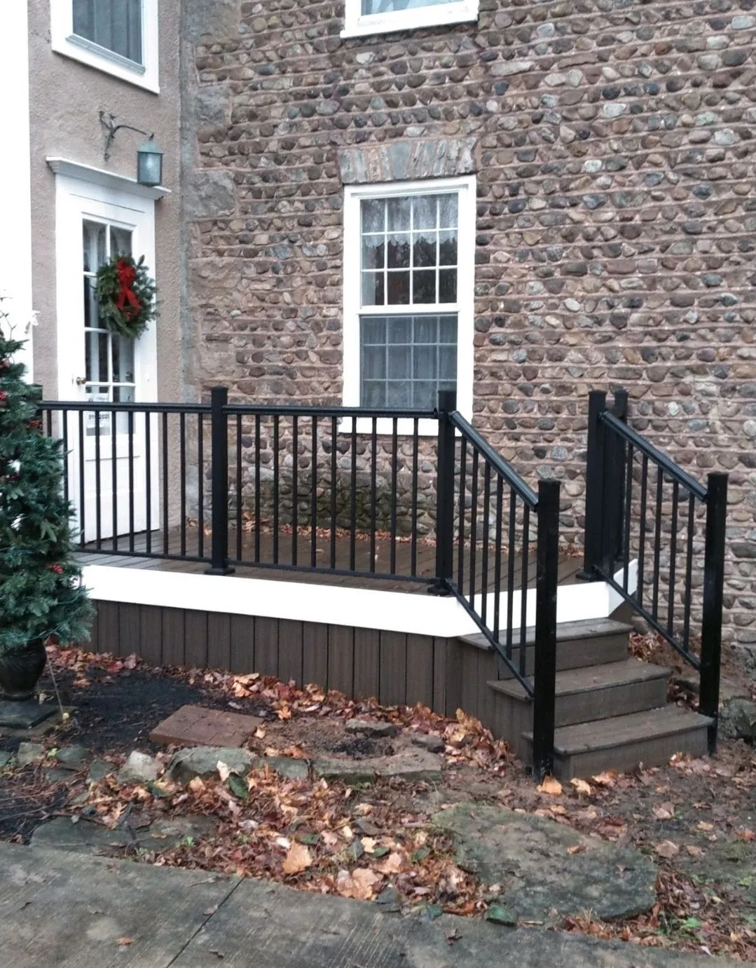 Black railing and stairs leading to a composite deck in front of a brick building; dried leaves on the ground.