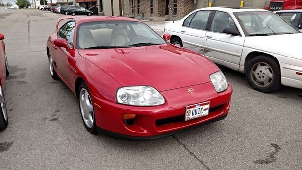 Red Toyota Supra parked on asphalt, facing forward. White car beside it. Buildings in the background.