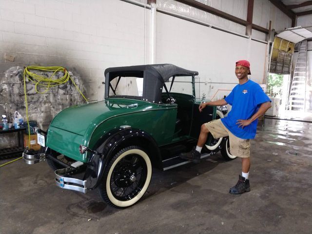 Man poses next to a classic green car with black wheels and a convertible top in a garage.