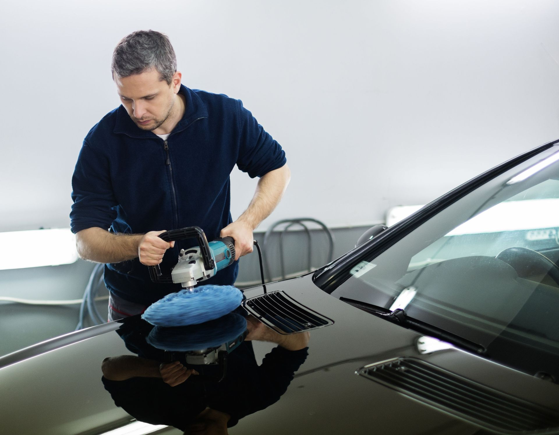 Man polishes the hood of a black car with a buffer in a garage.