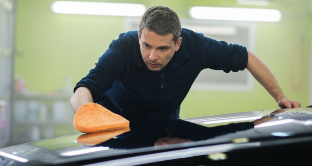 Man cleaning a black car’s hood with an orange cloth in a bright garage