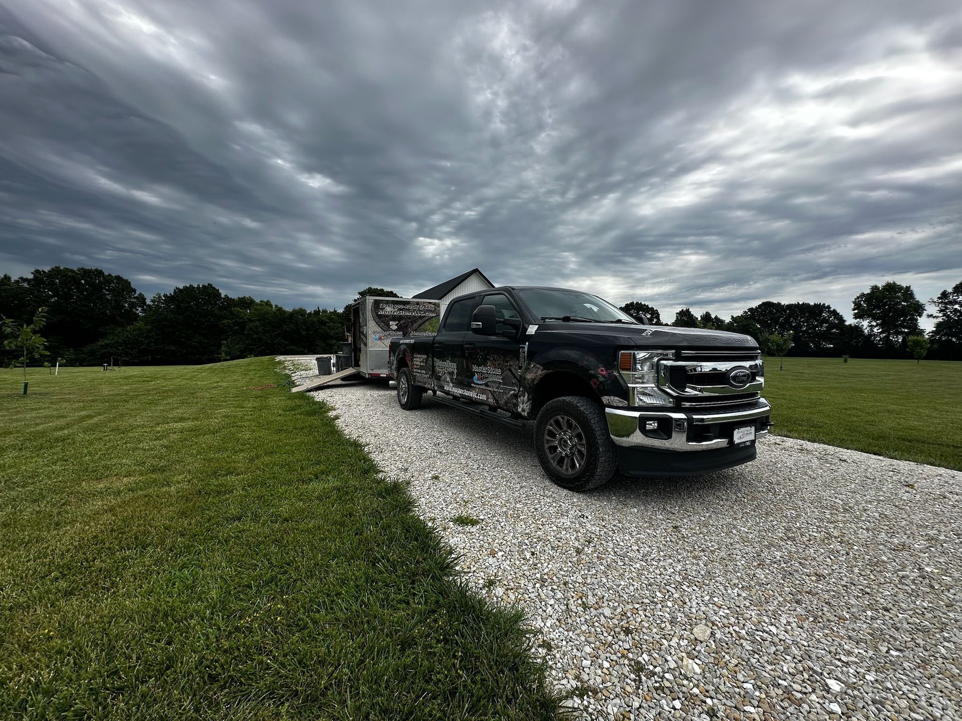 Black pickup truck towing a trailer on a gravel driveway beside a grassy field under cloudy skies