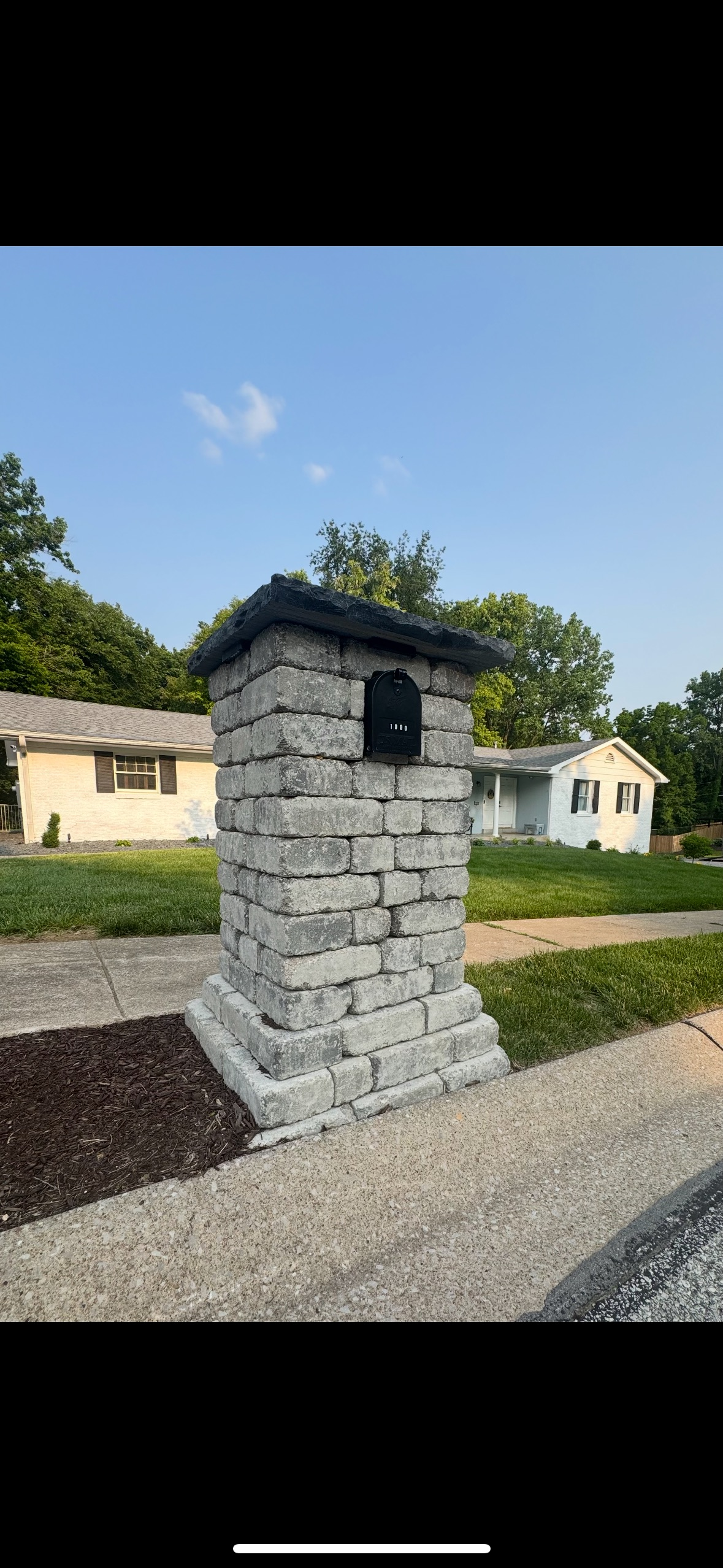 Stone mailbox pillar beside a driveway in a rural neighborhood under a blue sky