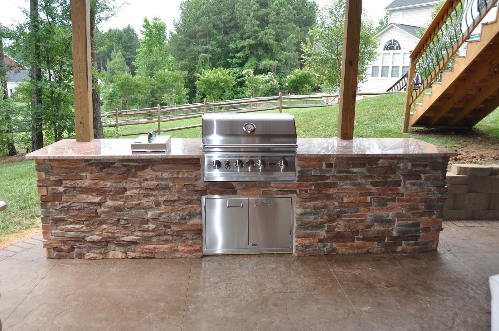 Outdoor stone grill island with stainless steel barbecue under a covered patio, overlooking a green yard