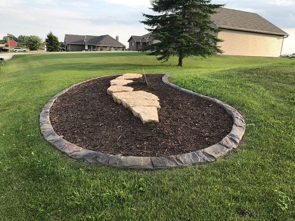Curved stone-bordered mulch bed with a tan rock centerpiece in a grassy suburban yard