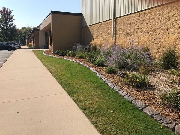 Sidewalk beside a tan commercial building with landscaped rocks, shrubs, and a grassy strip