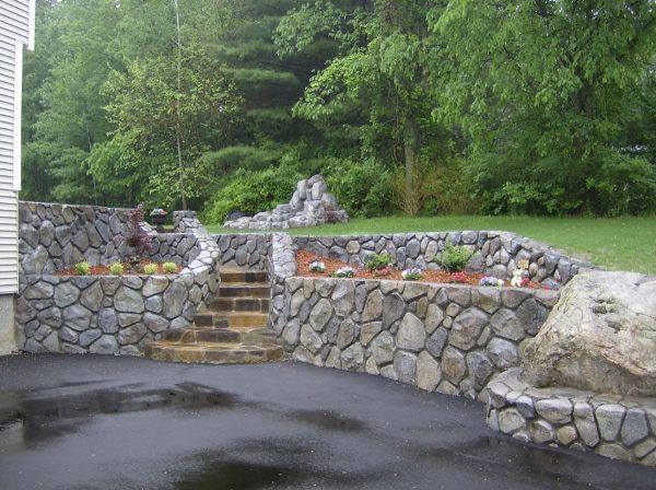 Stone retaining walls and steps in a landscaped backyard with trees and a wet driveway