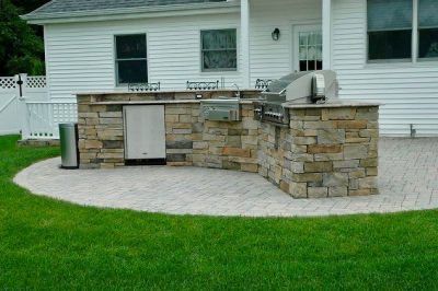 Backyard stone outdoor kitchen with grill against a white house, beside a paved patio and green lawn