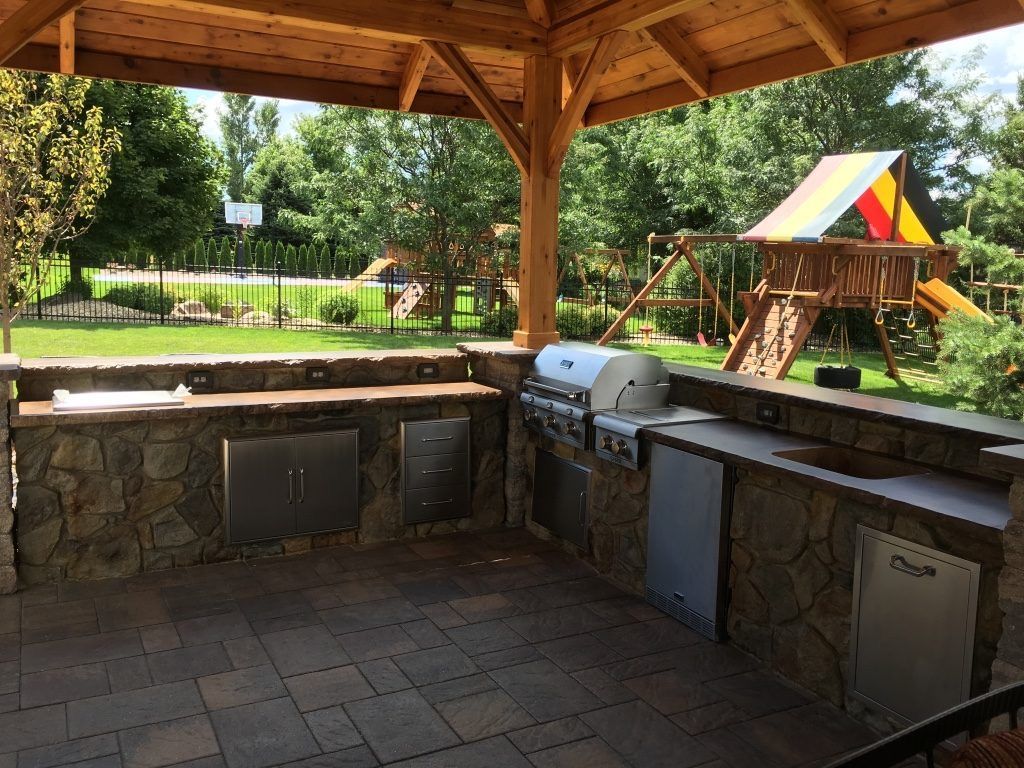 Outdoor stone grill area under a wooden pavilion with a grassy yard and colorful playground in the background