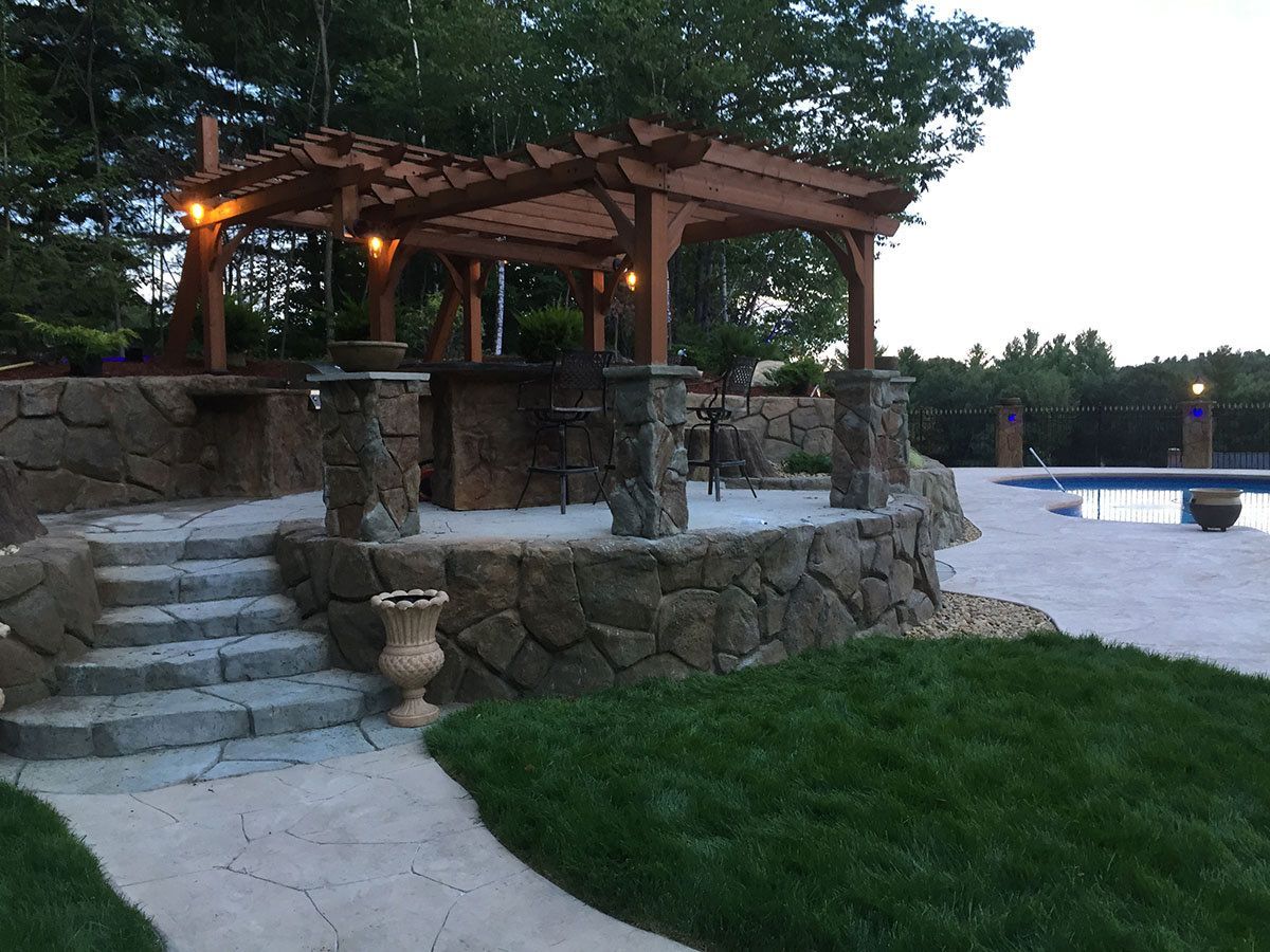 Stone pavilion with wooden roof by a pool, stone steps, and green lawn at dusk
