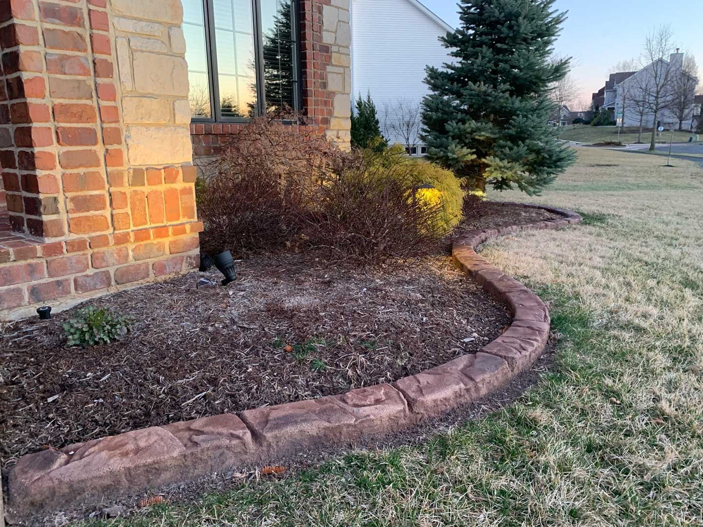 Curved brick garden bed beside a house with shrubs, a small evergreen, and frost-dusted grass