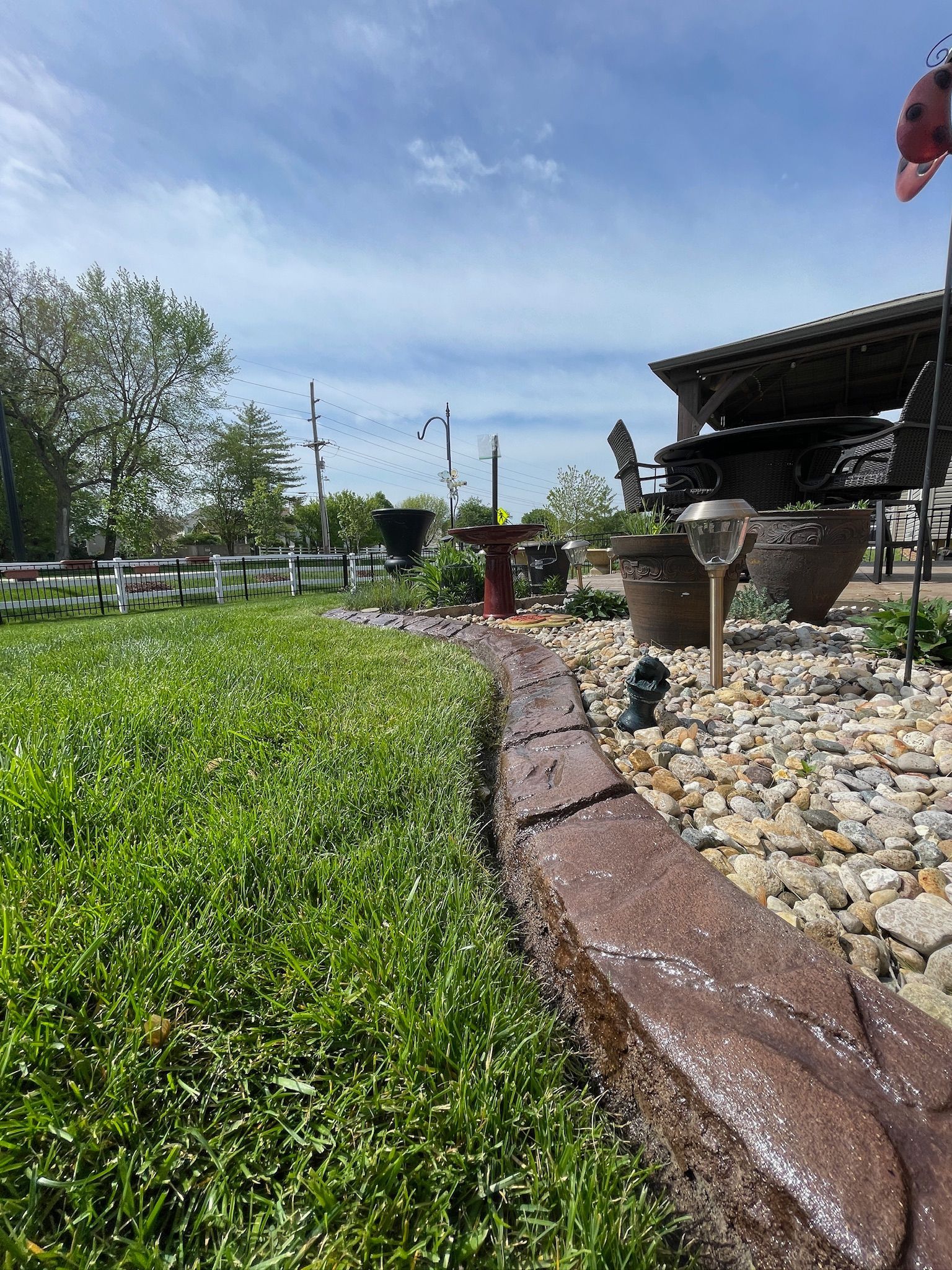 Sunny garden with green lawn, stone border, and a bird feeder beside a wooden structure.