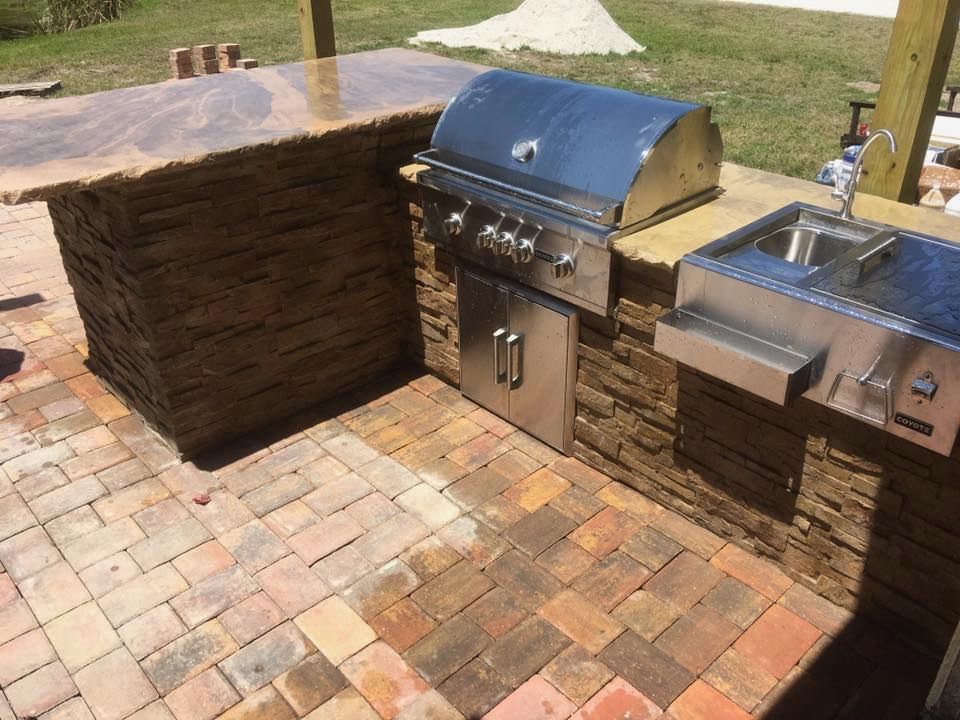 Outdoor built-in grill and sink with stone countertops on a brick patio in a backyard