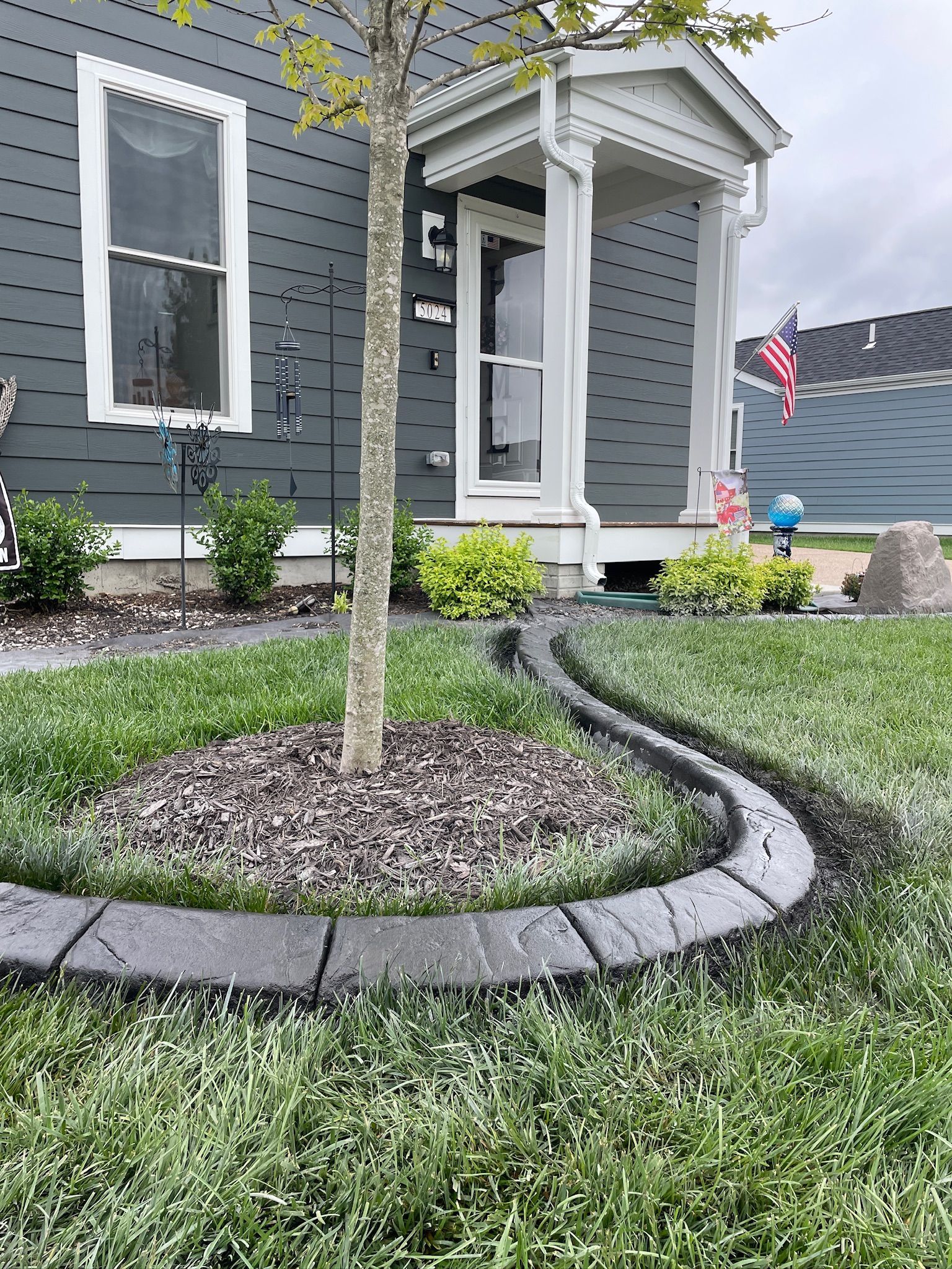 Front yard with a young tree, curved stone edging, and a gray house porch with an American flag.