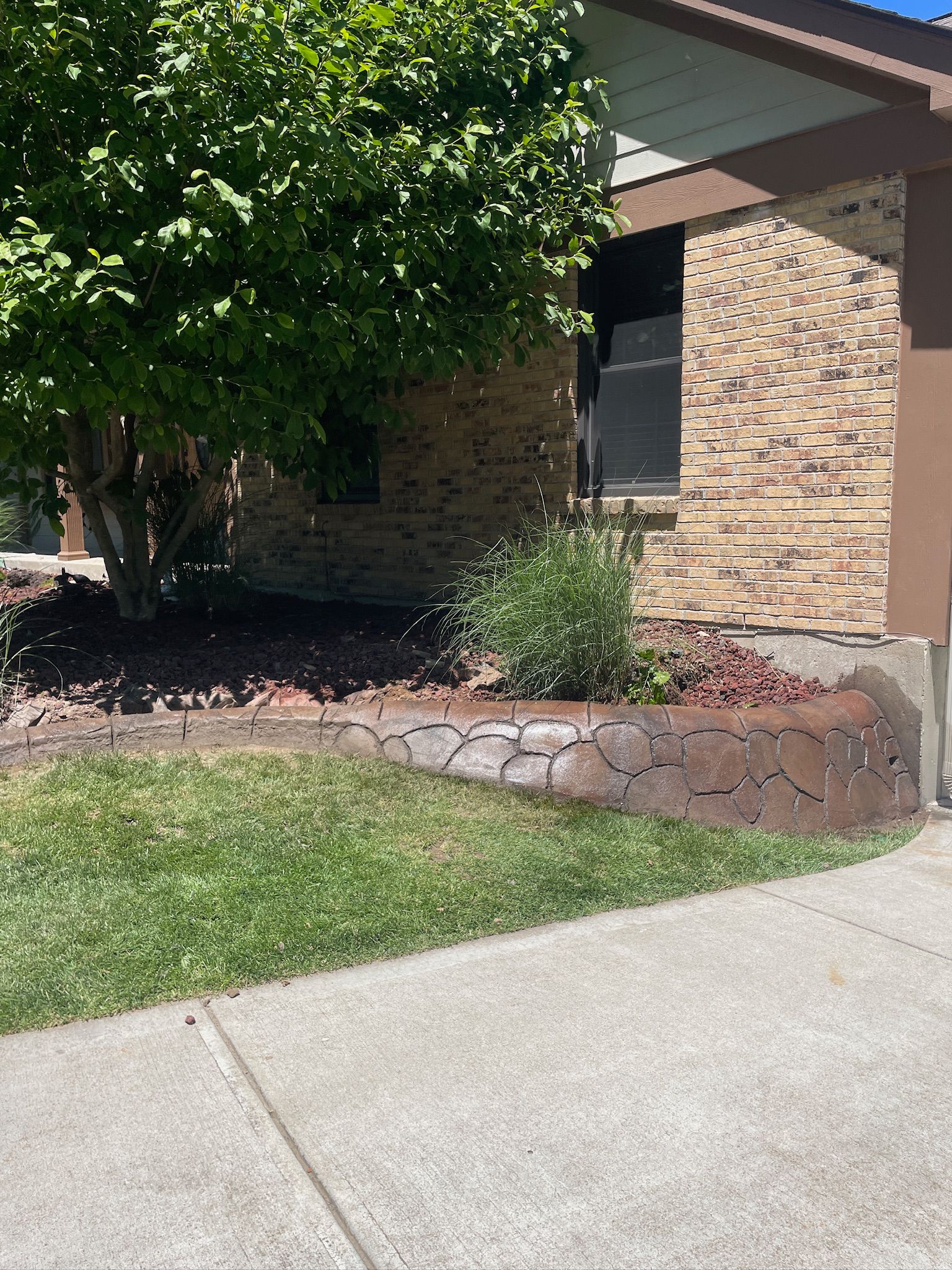 Brick house front yard with a tree, mulch bed, grass lawn, and concrete walkway.