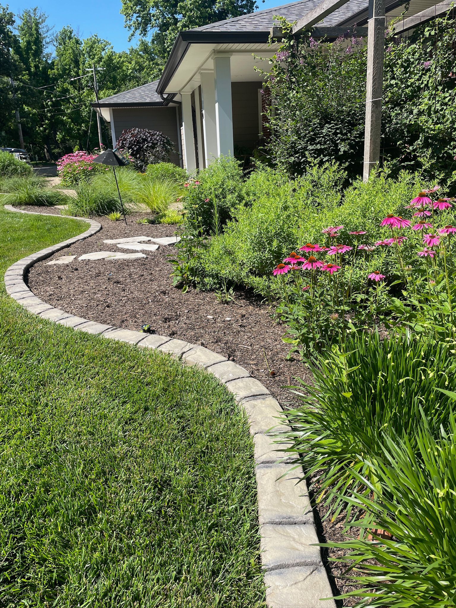 Curved stone path through a lush garden beside a house, with pink flowers and greenery in bright sunlight