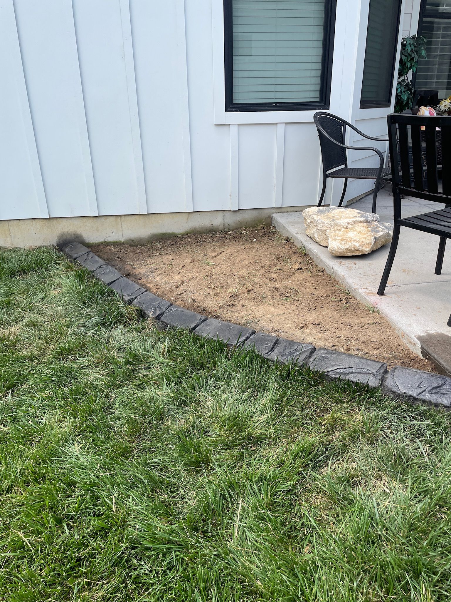 Small landscaped mulch bed bordered by stones beside a house and patio chairs, with grass in the foreground