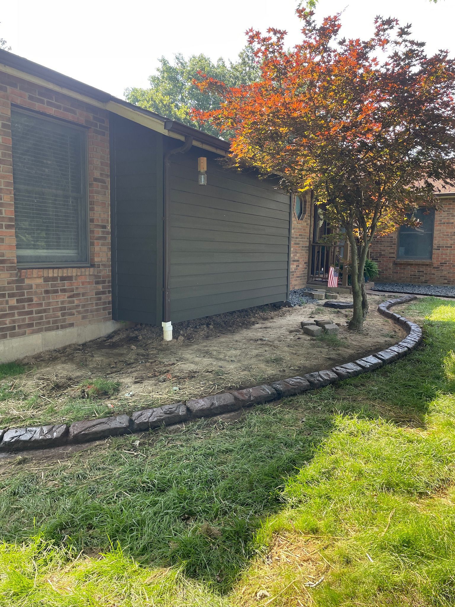 Single-story brick house with a gray extension, autumn tree, and curved stone border along the lawn
