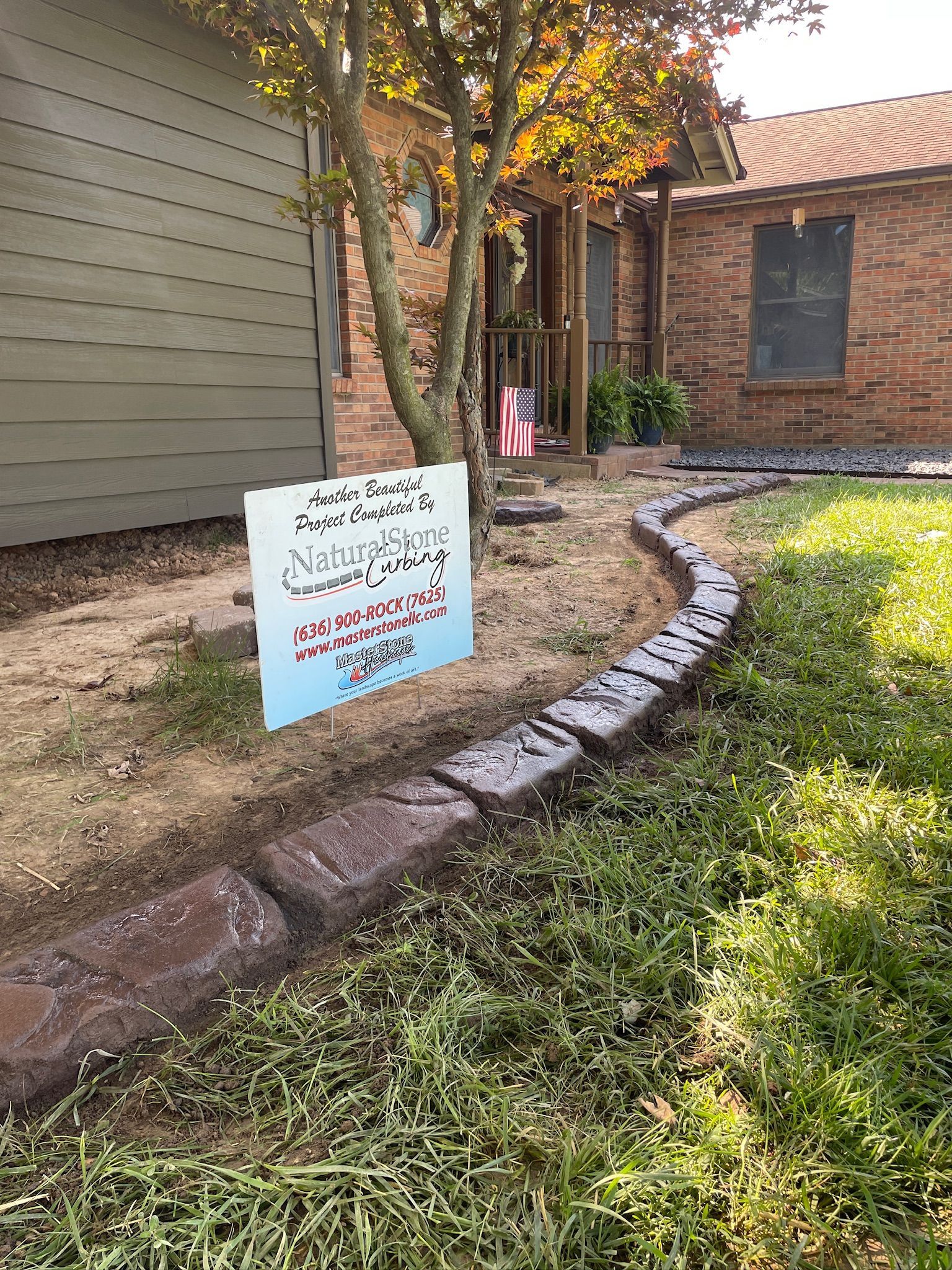 Yard sign beside a brick house with Arabic text and pink flowers along a curved border