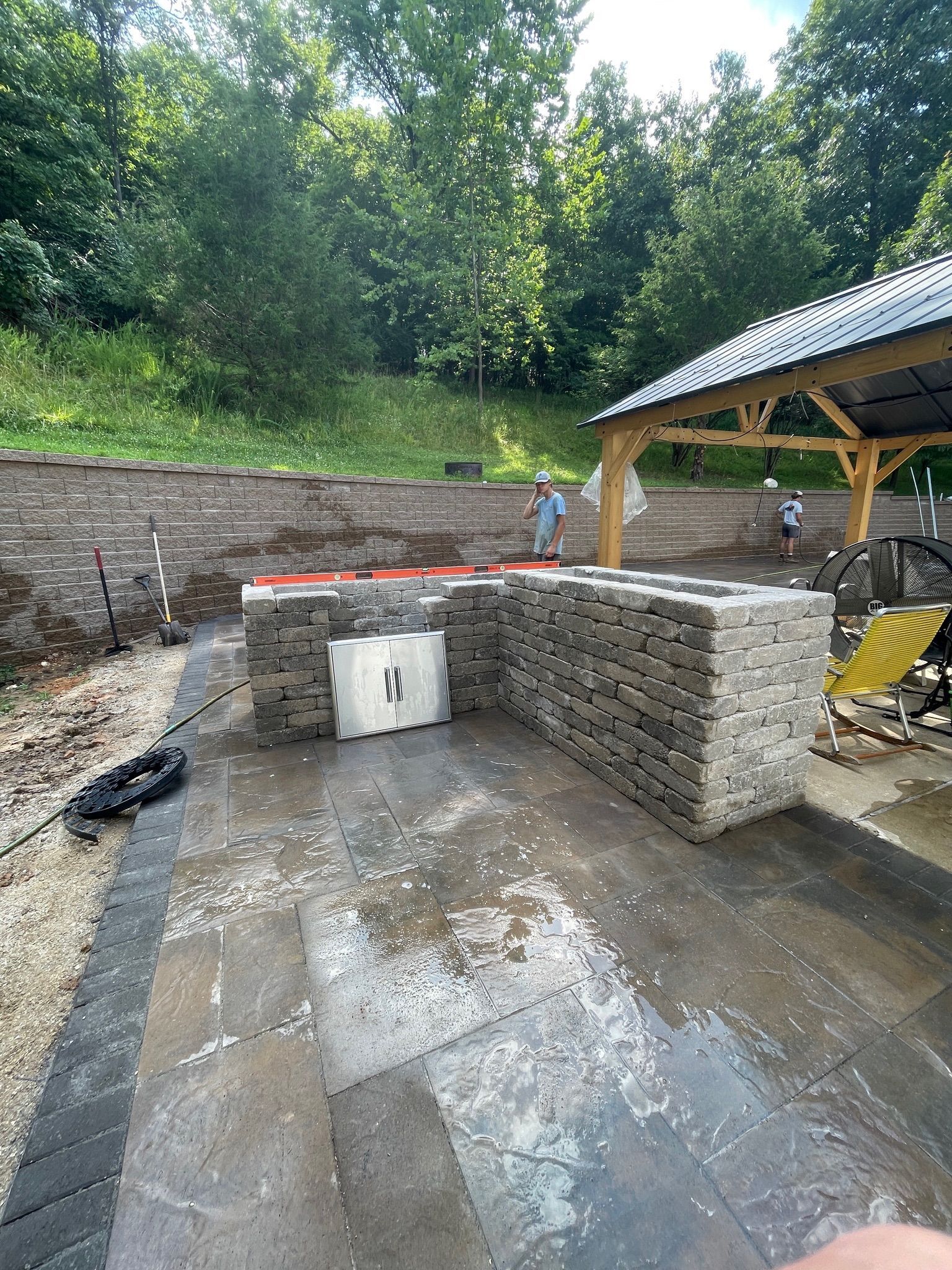 Backyard stone retaining walls around a wet concrete pad beside a pavilion and wooded slope