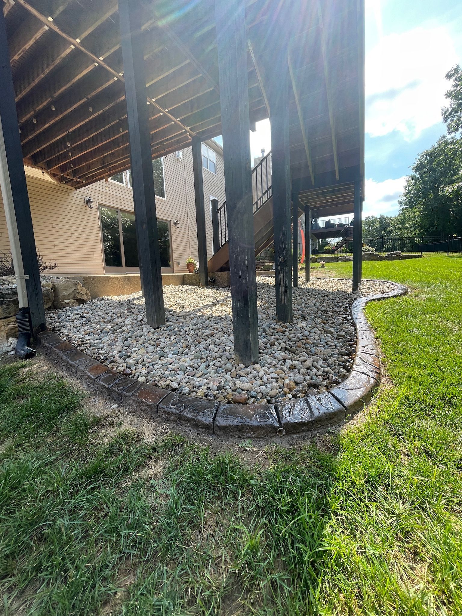 Under a raised wooden deck, black posts stand in a stone-bordered gravel bed beside grass.