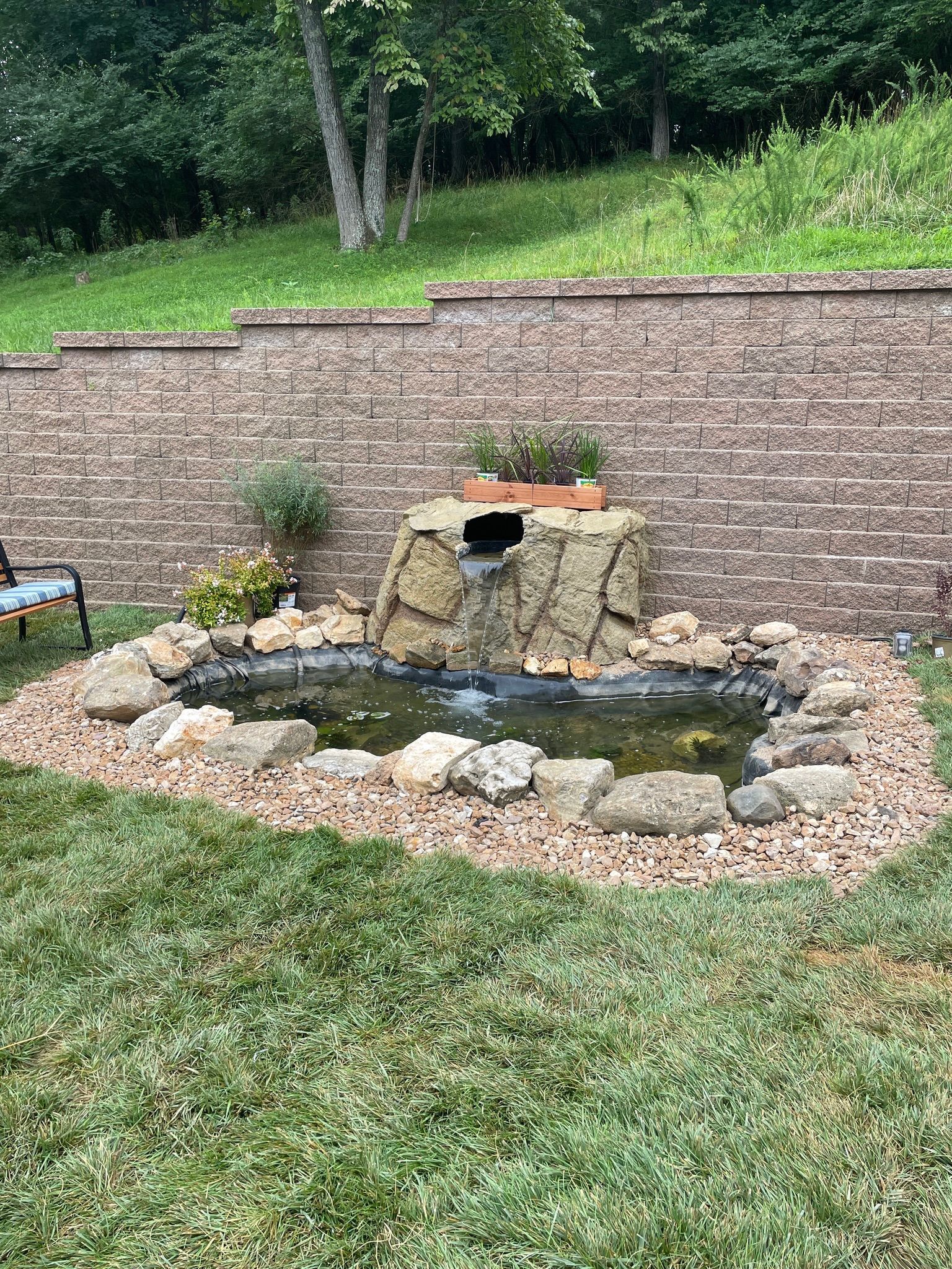 Small backyard pond with a stone waterfall, rock border, and brick retaining wall behind it