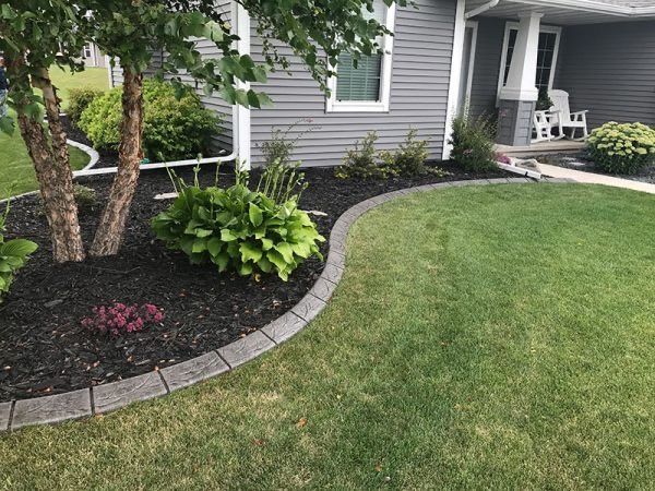 Curved stone garden bed with shrubs and mulch beside a gray house and green lawn