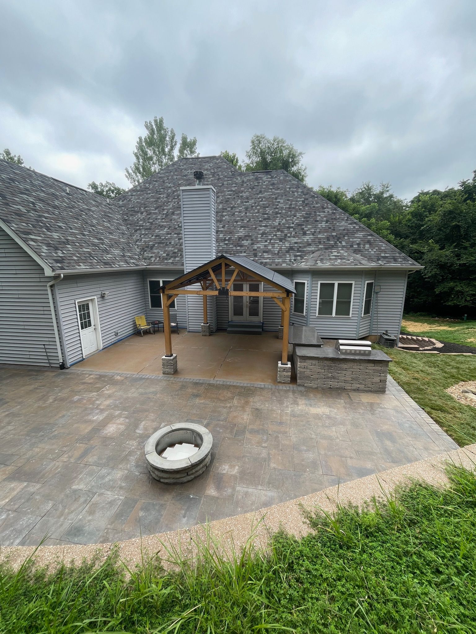 Backyard patio with wooden pergola, stone fire pit, and a house exterior under cloudy sky