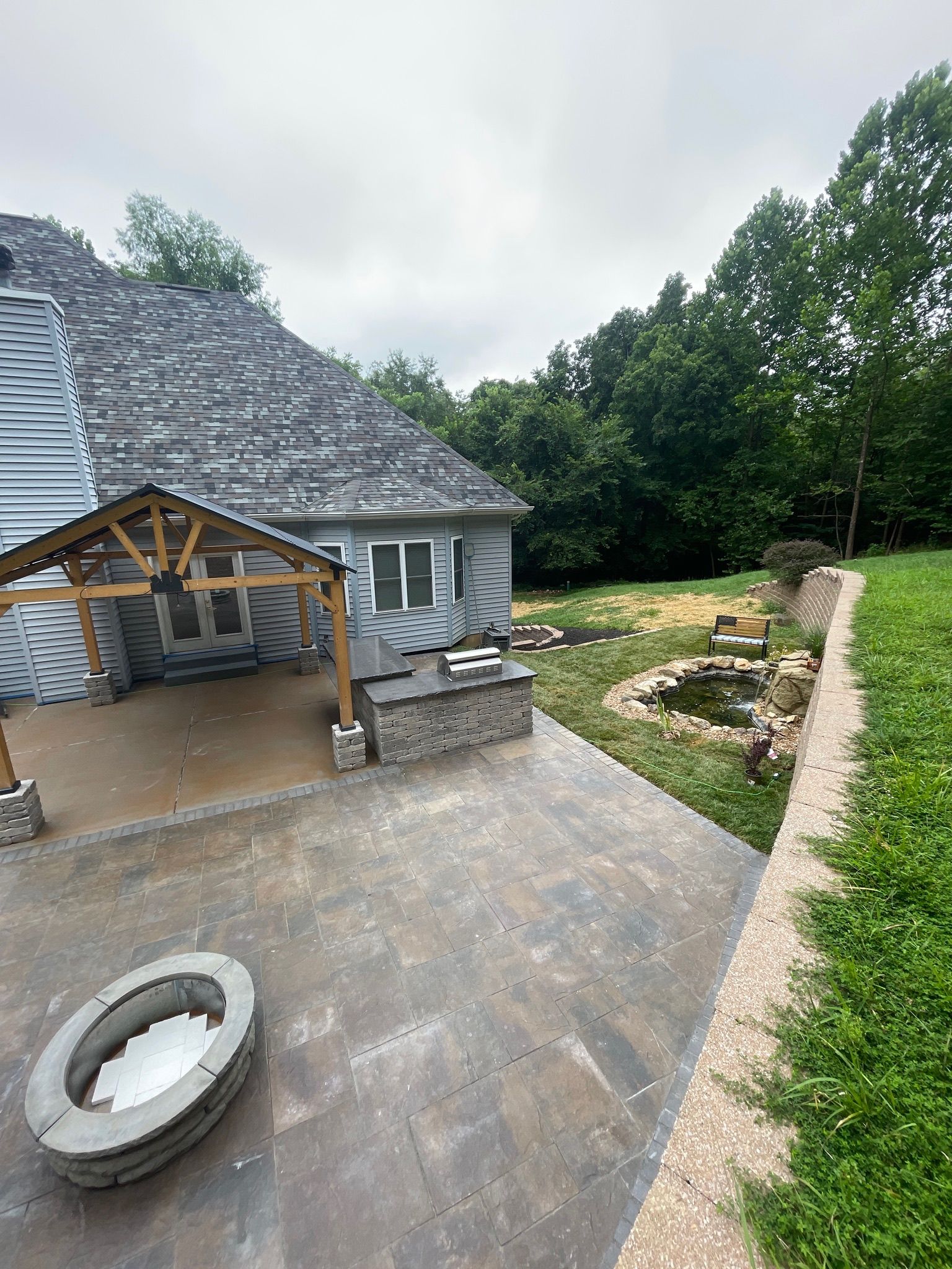 House patio with stone paving, a covered seating area, and a landscaped garden beside a wooded yard