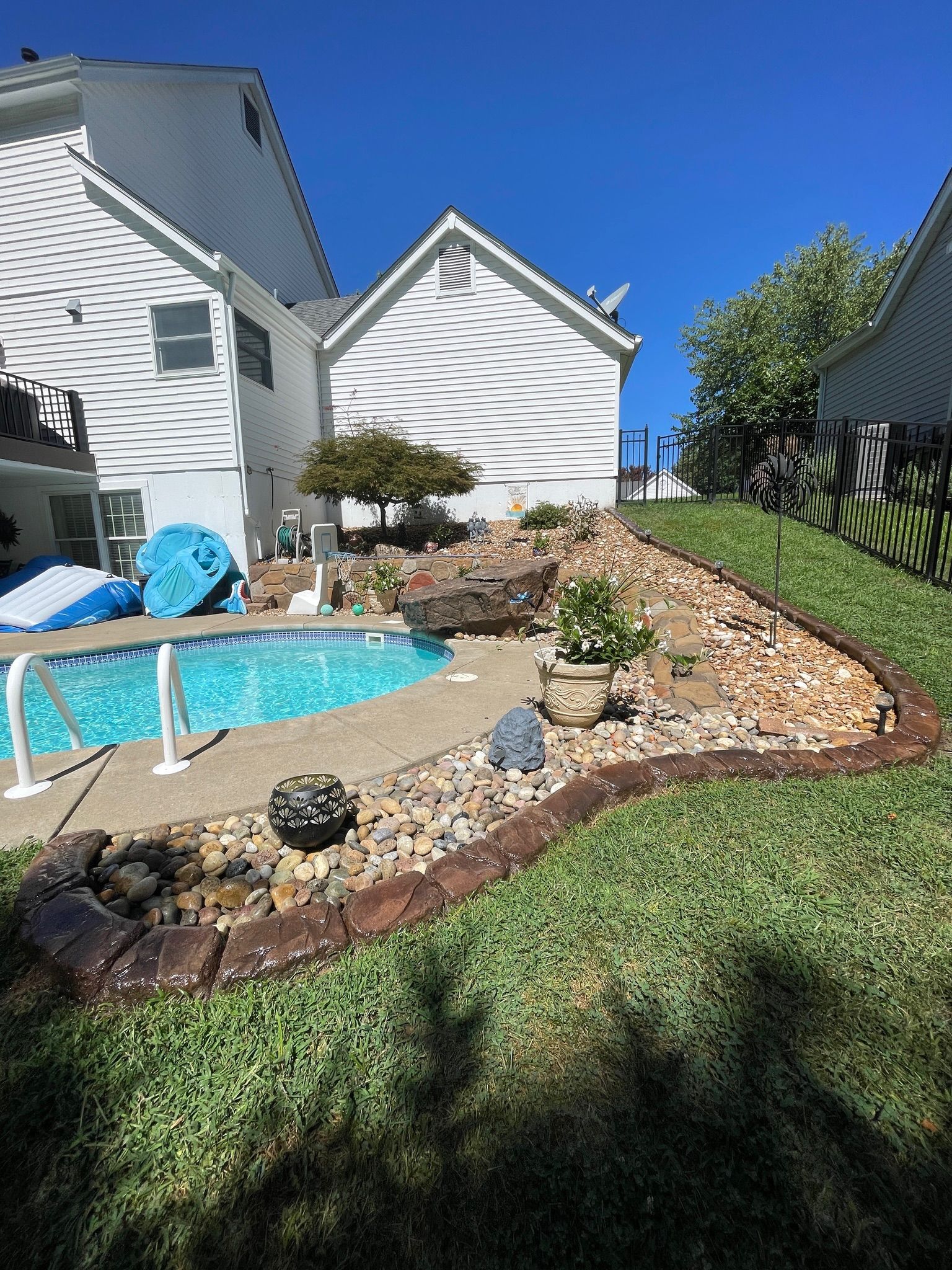 Backyard with a small pool, stone landscaping, green lawn, and white house under a blue sky