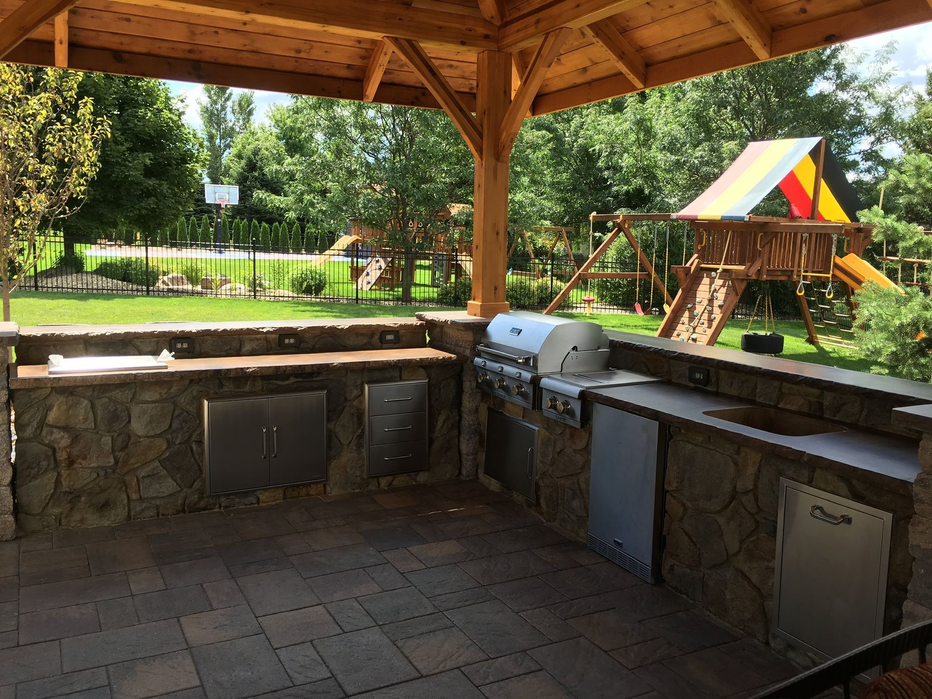 Covered outdoor stone grilling area with two grills and a colorful play structure in the background