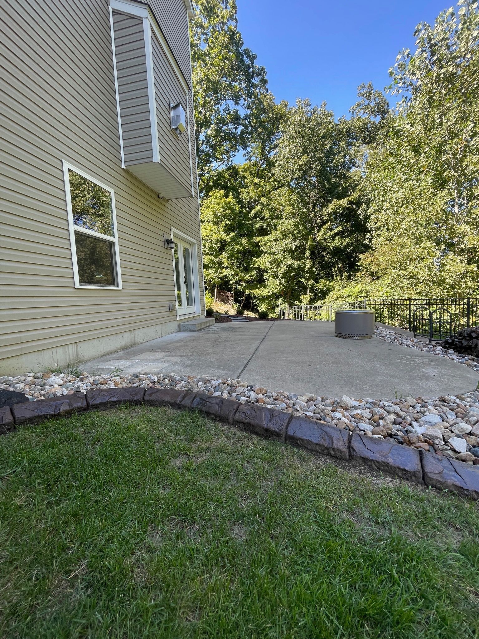 Backyard patio beside beige house, with stone border, grass, trees, and a small planter.