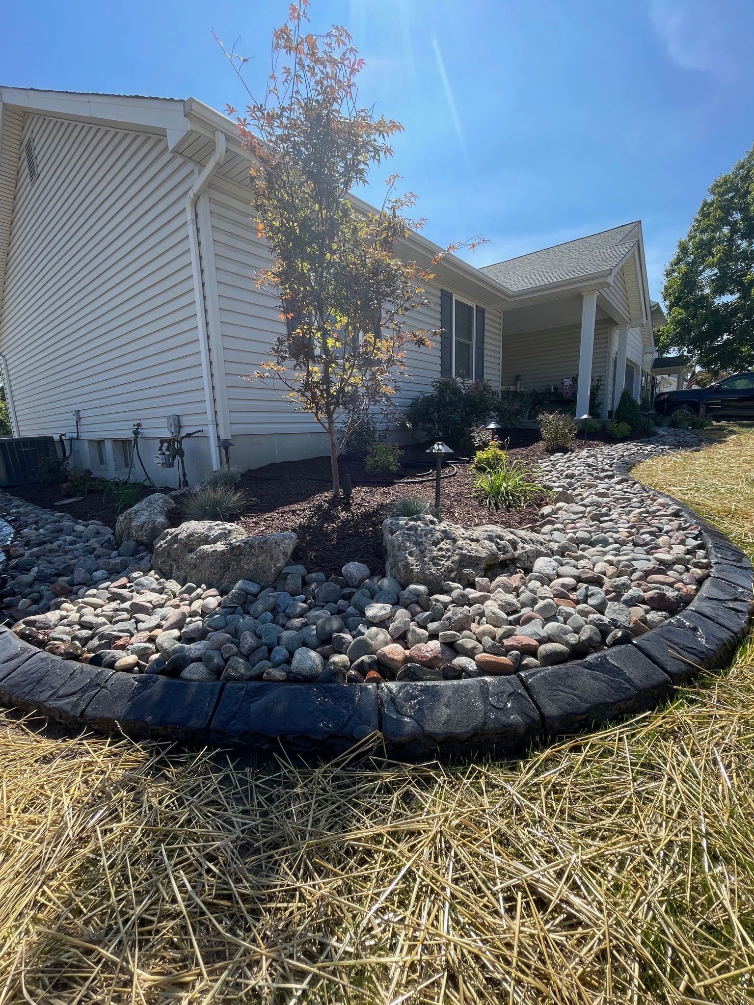 Landscaped front yard with white house, young tree, and circular stone-bordered garden bed under bright sun