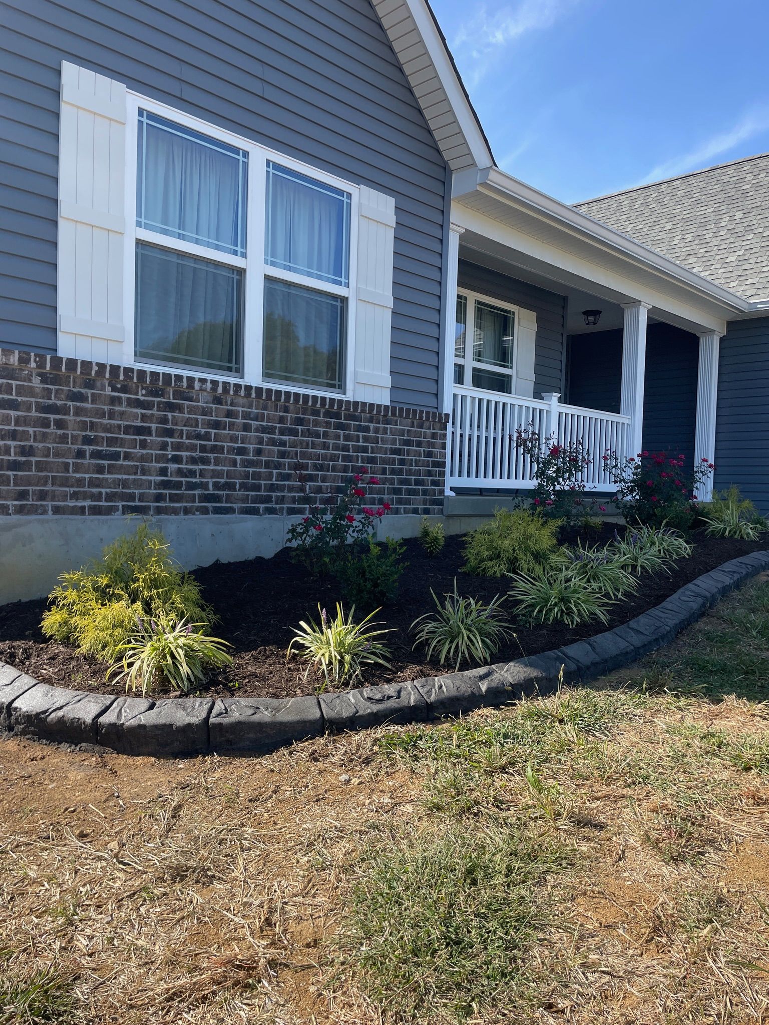 Brick house exterior with white shutters, front porch, and landscaped shrubs along the foundation.