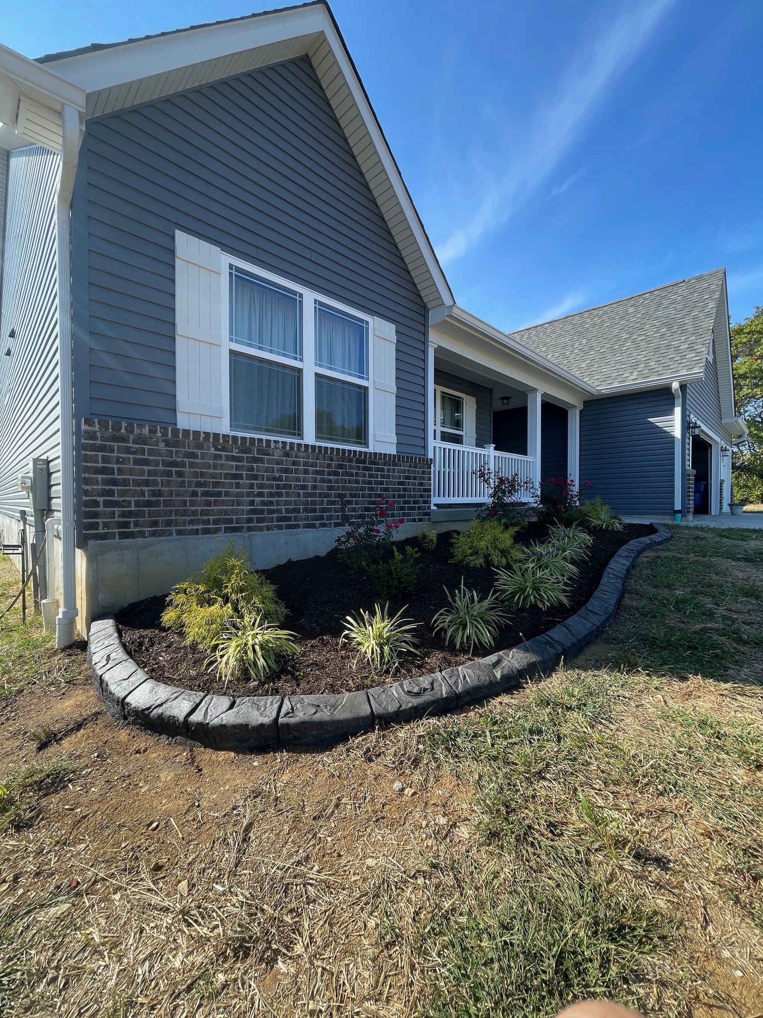 Front of a gray house with white trim, brick accents, and a curved landscaped flower bed under a blue sky