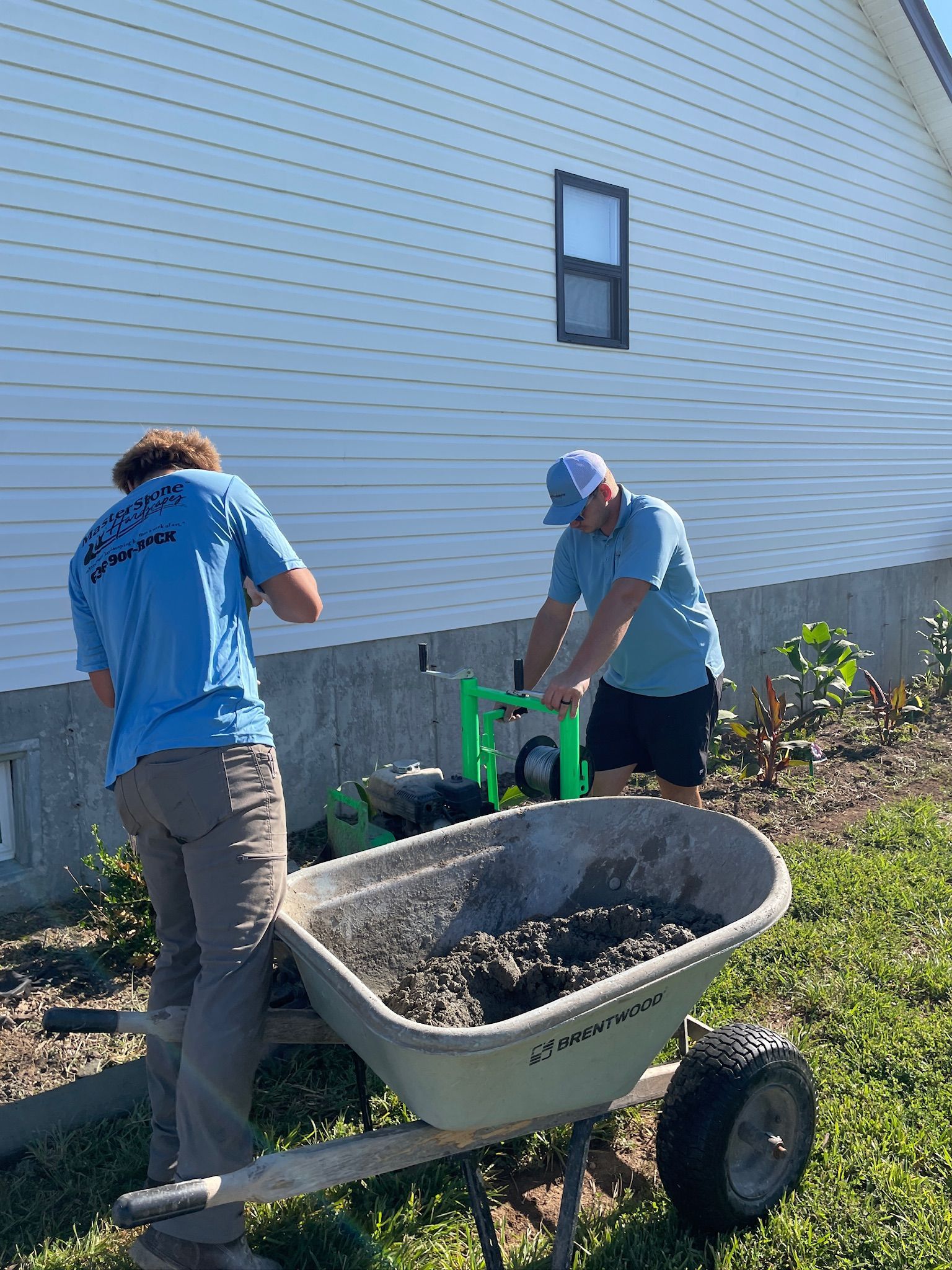 Two people push a wheelbarrow of soil beside a house, with a green compost bin nearby.