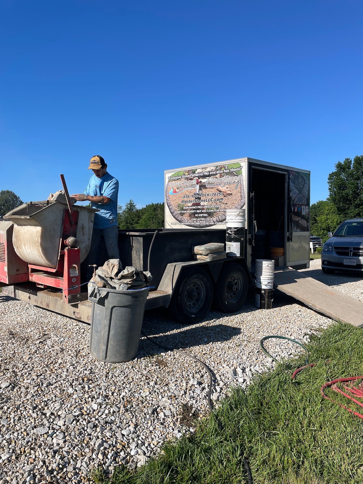 Worker on a truck-mounted lift beside a gravel driveway, with a red dumpster and blue sky.
